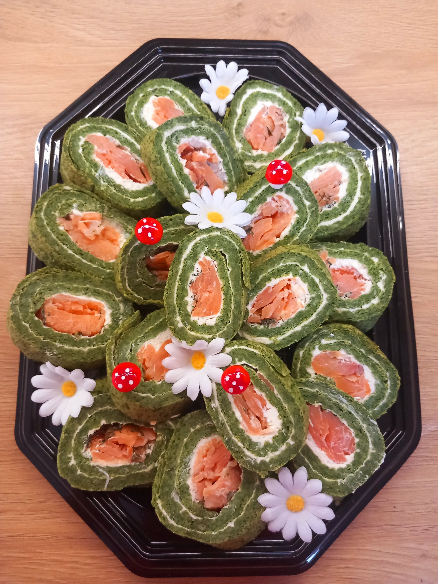 Dekoracyjnie ułożone roladki szpinakowe z łososiem i kremowym serem na czarnej tacy, ozdobione małymi, sztucznymi kwiatkami i grzybkami.