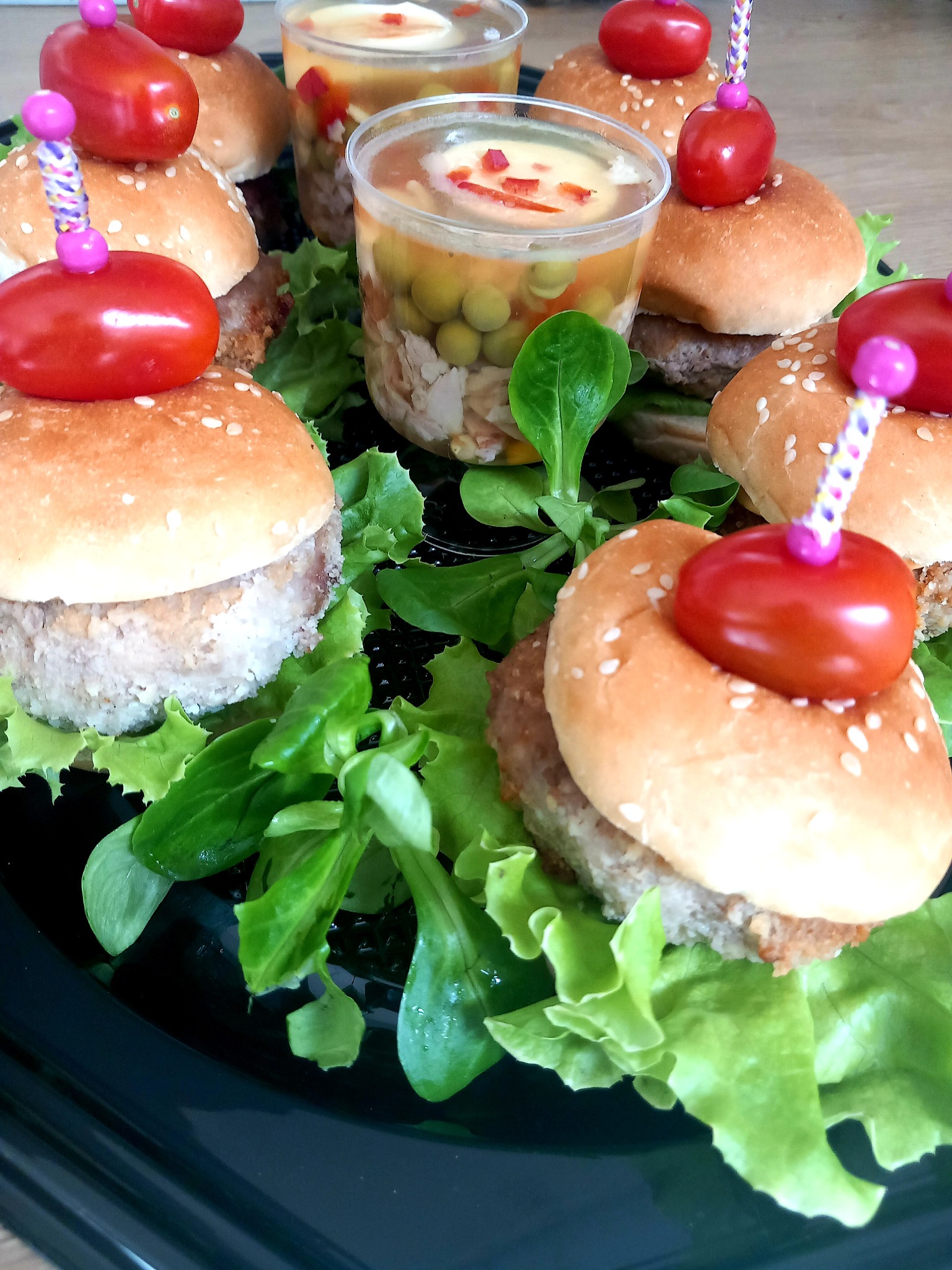 Miniaturowe burgery z sezamem i pomidorkami koktajlowymi na wykałaczkach, obok galaretki warzywnej w plastikowych kubeczkach, ułożone na sałacie polnej na ciemnym talerzu.