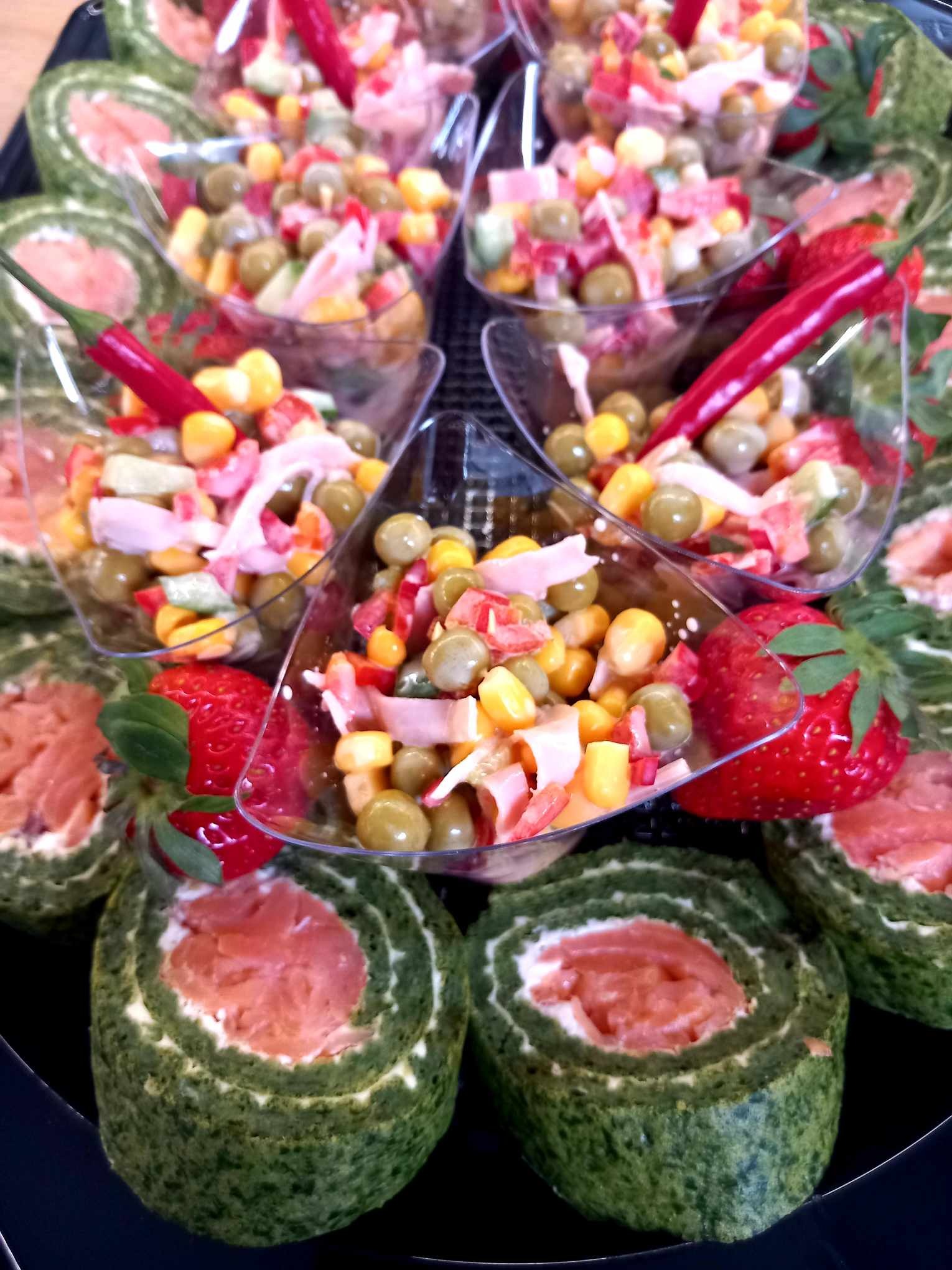 Dekoracyjna kompozycja cateringowa: roladki szpinakowe z łososiem, sałatka warzywna w trójkątnych miseczkach, ozdobiona truskawkami i czerwoną papryczką chili.