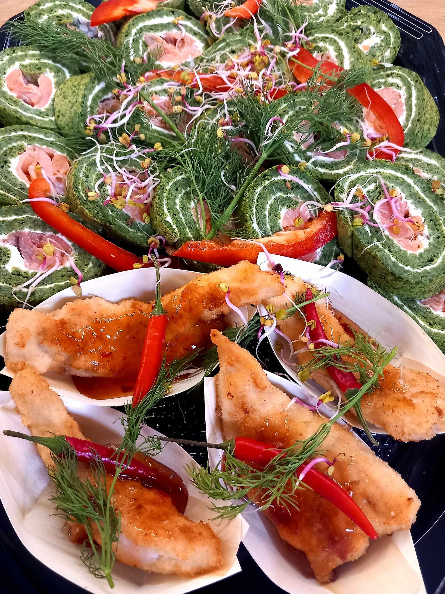 Elegancko podane przekąski: rolady szpinakowe z łososiem i smażone ryby w sosie słodko-pikantnym, udekorowane świeżym koperkiem, kiełkami i papryczkami chili, na czarnym talerzu.