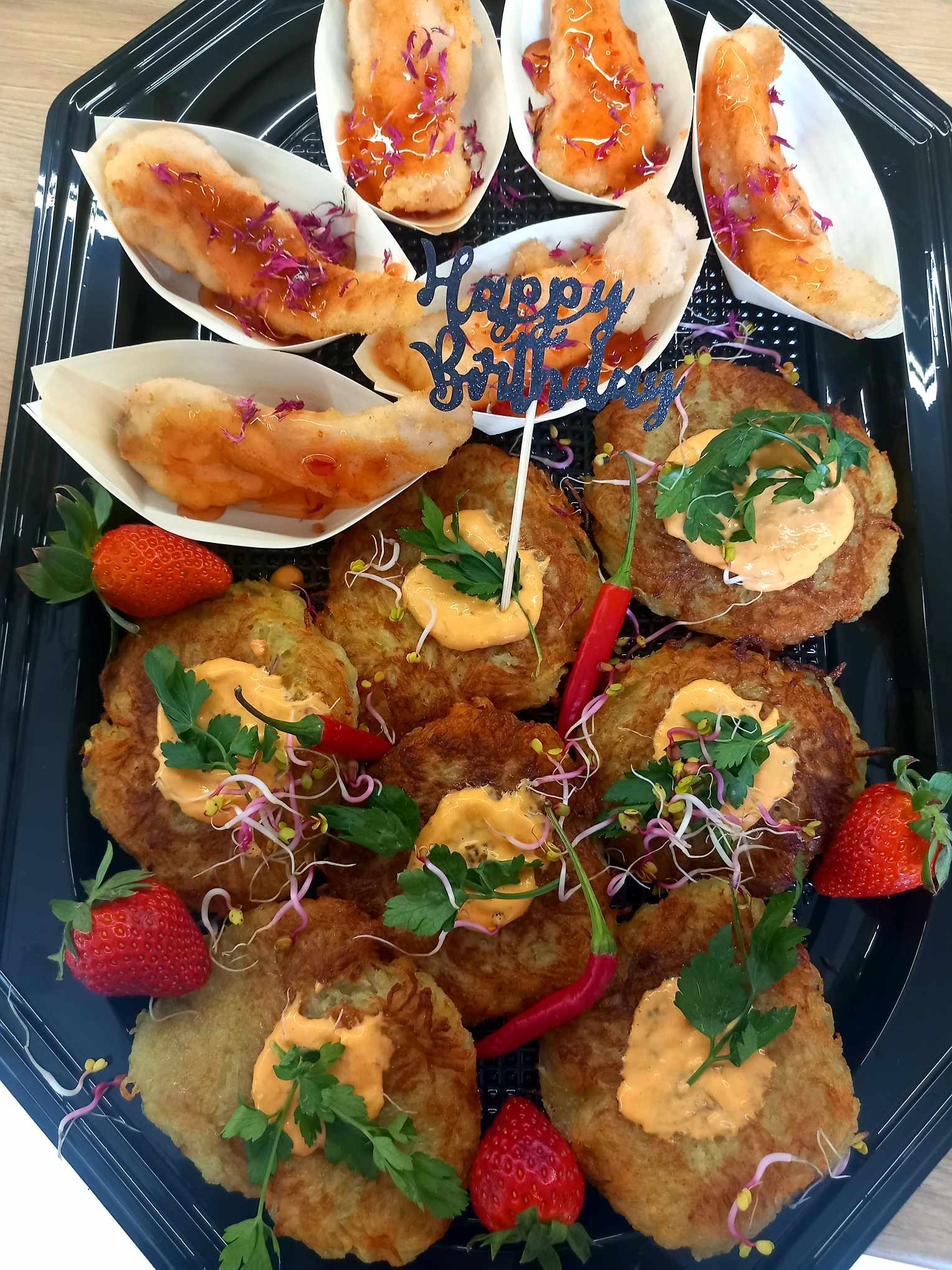 Czarny, plastikowy talerz z ziemniaczanymi plackami z sosem, udekorowanymi natką pietruszki, kiełkami i czerwoną papryczką chili, obok truskawki, a w tle łódeczki z rybą w sosie i dekoracją...