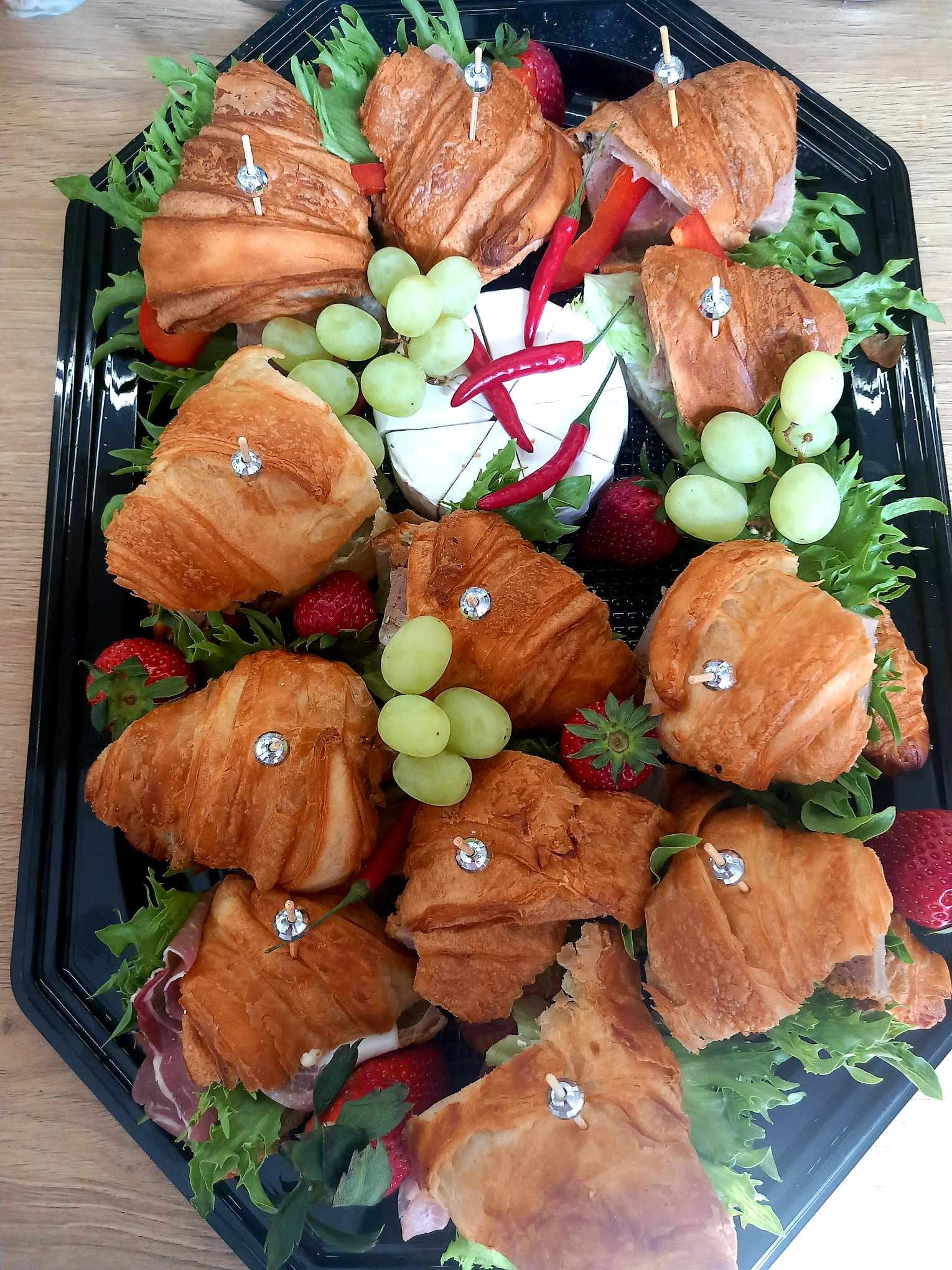 Czarny cateringowy półmisek z mini-croissantami z szynką, serem brie, winogronami, truskawkami i papryczkami chili, udekorowany zielonymi liśćmi sałaty.