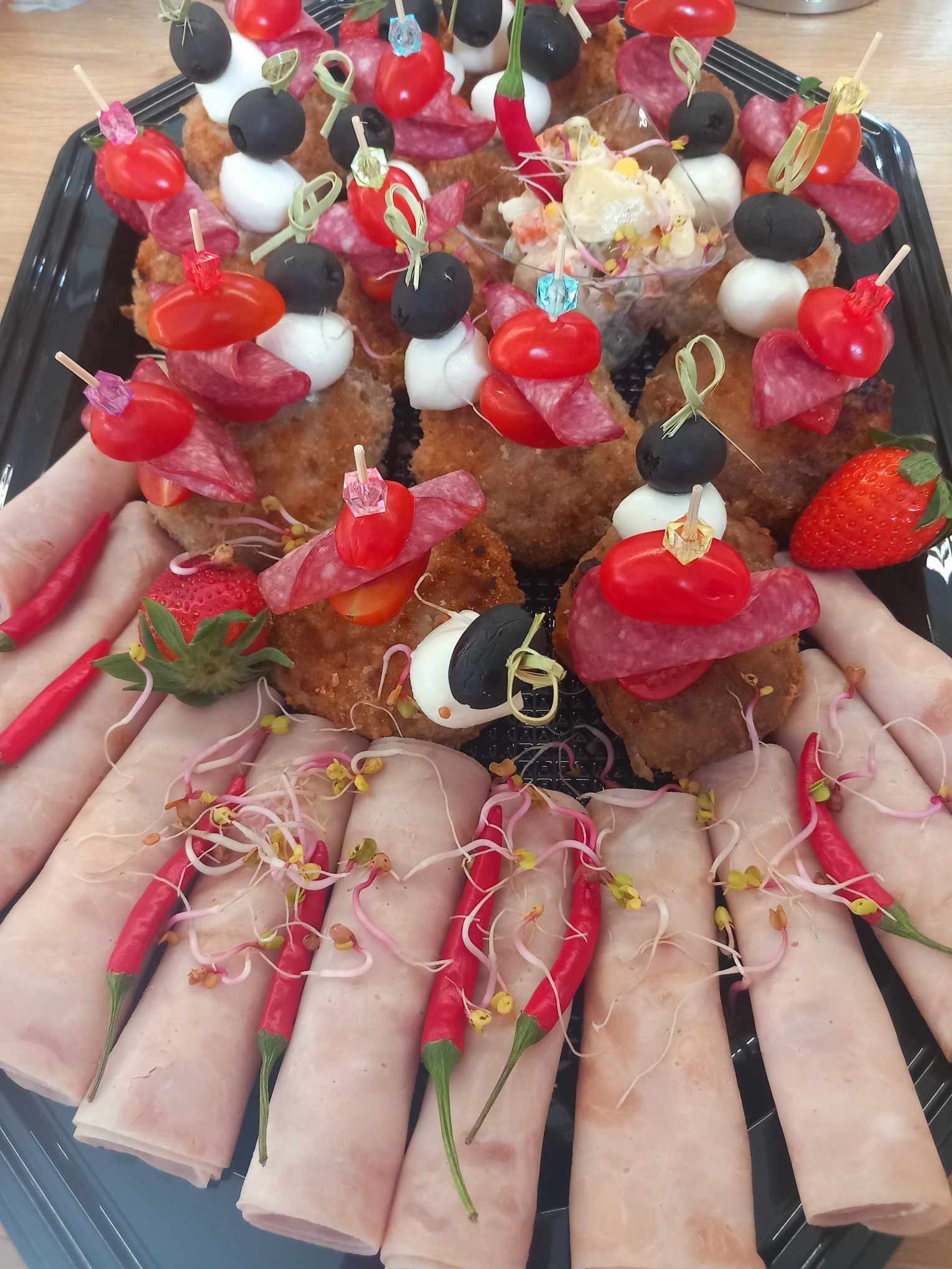 Czarny cateringowy półmisek z roladkami z szynki dekorowanymi kiełkami, mini szaszłykami z salami, pomidorkami, mozzarellą i oliwkami oraz sałatką w przezroczystym naczyniu, całość uzupełniona...