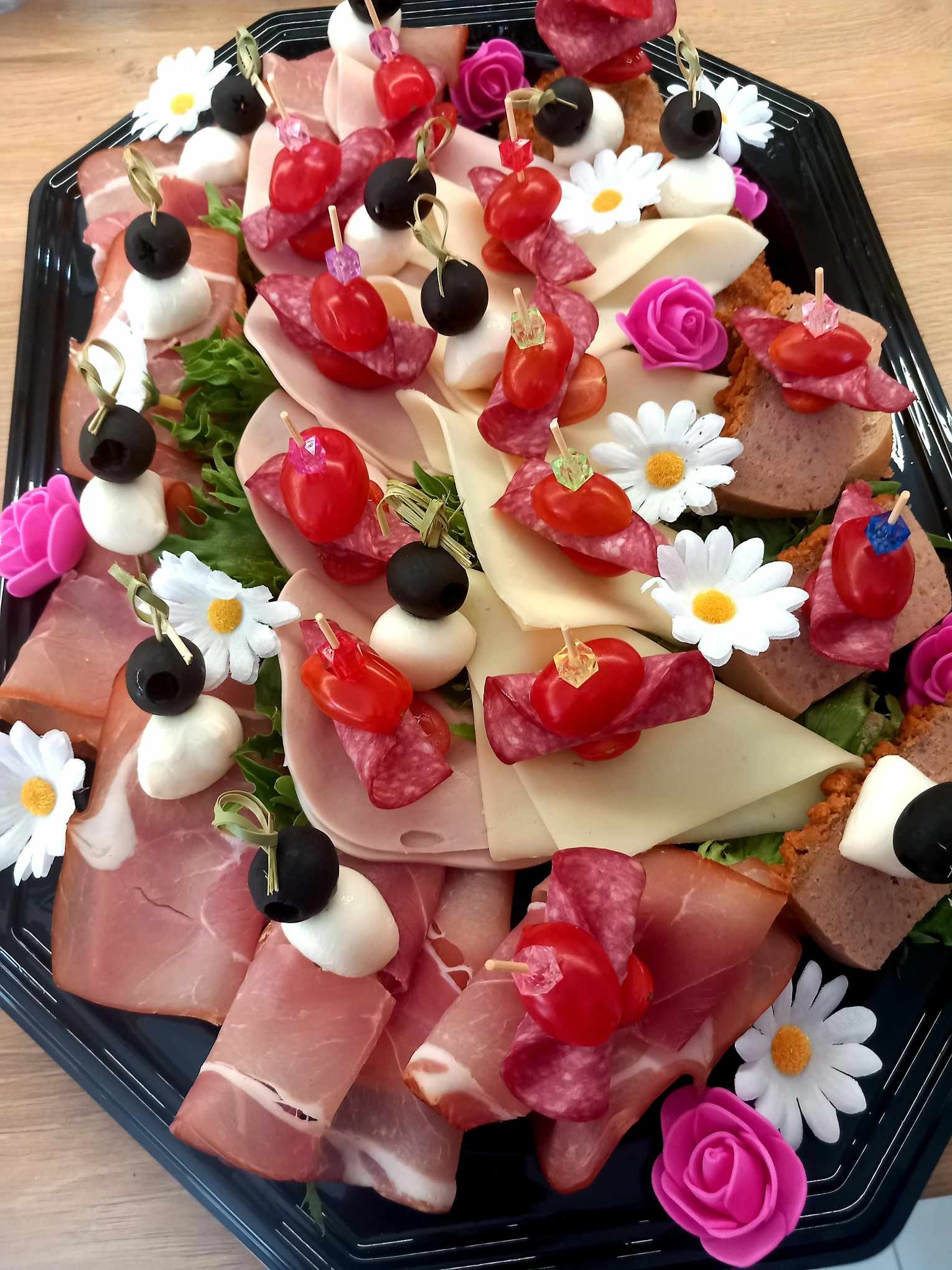 Dekoracyjna taca z przekąskami: koreczki z szynki parmeńskiej, salami, sera, pomidorków cherry i oliwek, ozdobiona sztucznymi kwiatami.