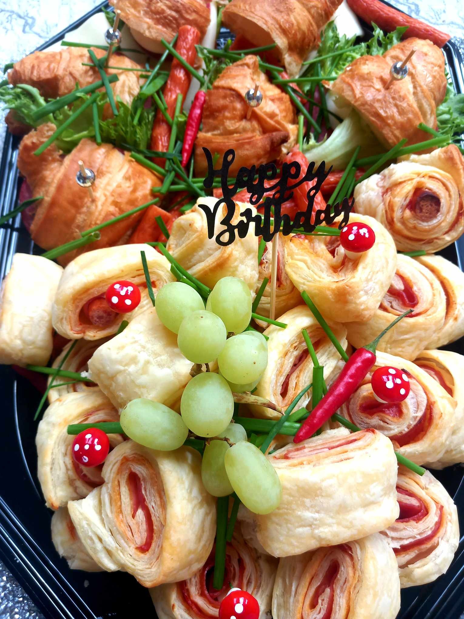 Dekoracyjna taca z przekąskami: roladki z ciasta francuskiego z szynką, mini croissanty z wkładką, zielony szczypiorek, winogrona i papryczki chili; czarny napis 'Happy Birthday'.