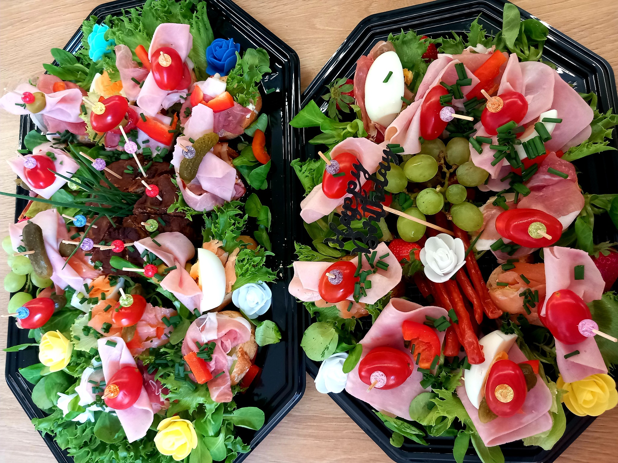 Dwie czarne tace z dekoracyjnymi przekąskami: kanapki z szynką, pomidorkami cherry i zieleniną, ozdobione małymi kwiatkami. Elegancki catering.