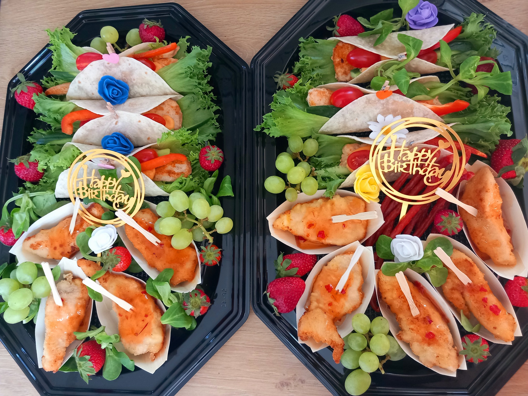 Dwie czarne tace z cateringiem: tortille z warzywami i mięsem, kawałki kurczaka w sosie, winogrona i truskawki. Dekoracje 'Happy Birthday'.