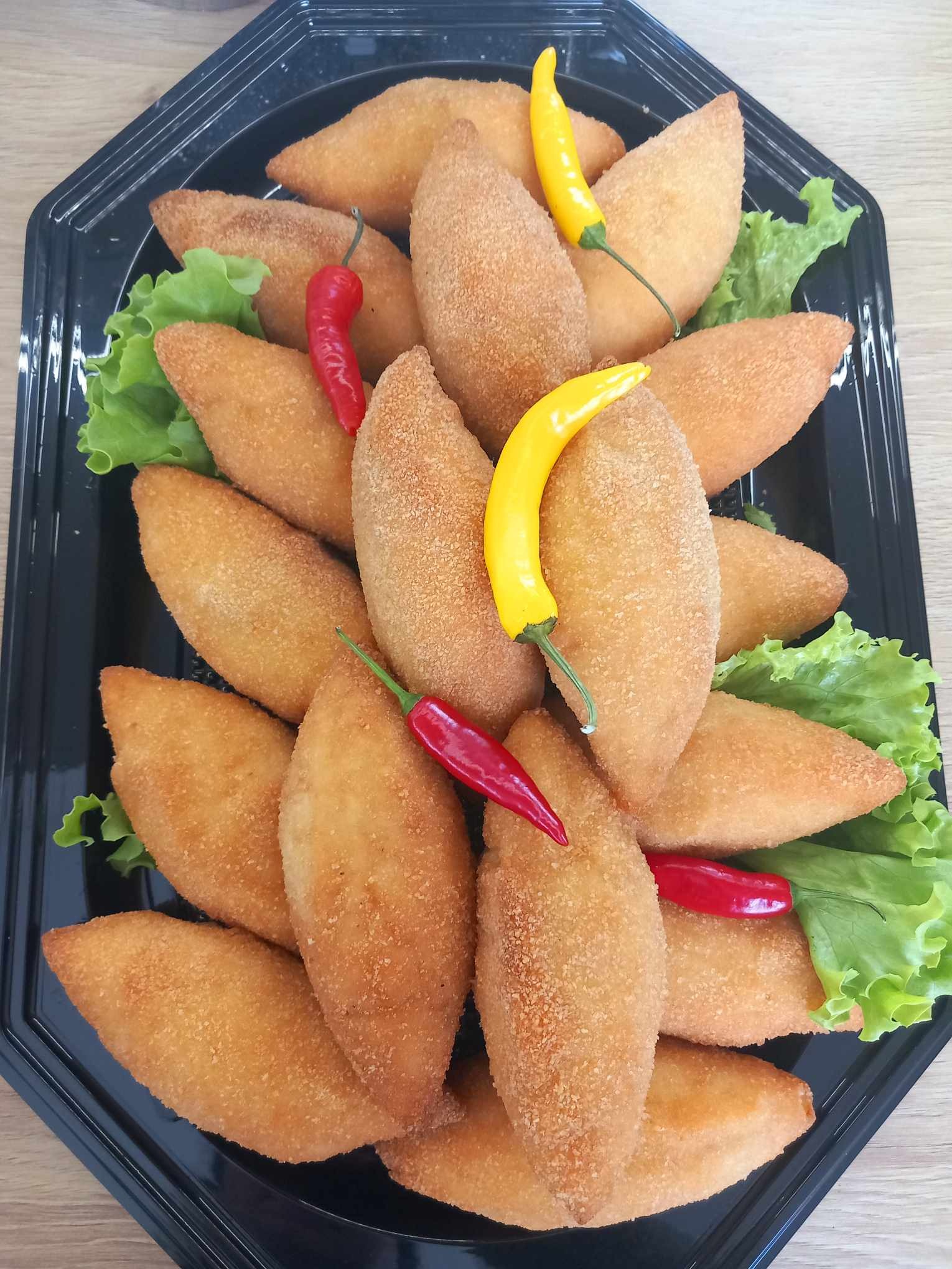 Czarna taca z krokietami, dekorowana sałatą i papryczkami chili (czerwona i żółta), prezentacja cateringu na stole z jasnego drewna.