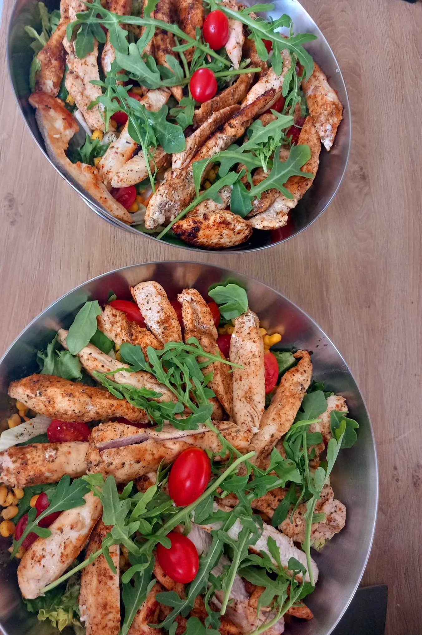 Dwie owalne miski ze stali nierdzewnej wypełnione sałatką z grillowanym kurczakiem, rukolą i pomidorkami koktajlowymi na drewnianym stole.