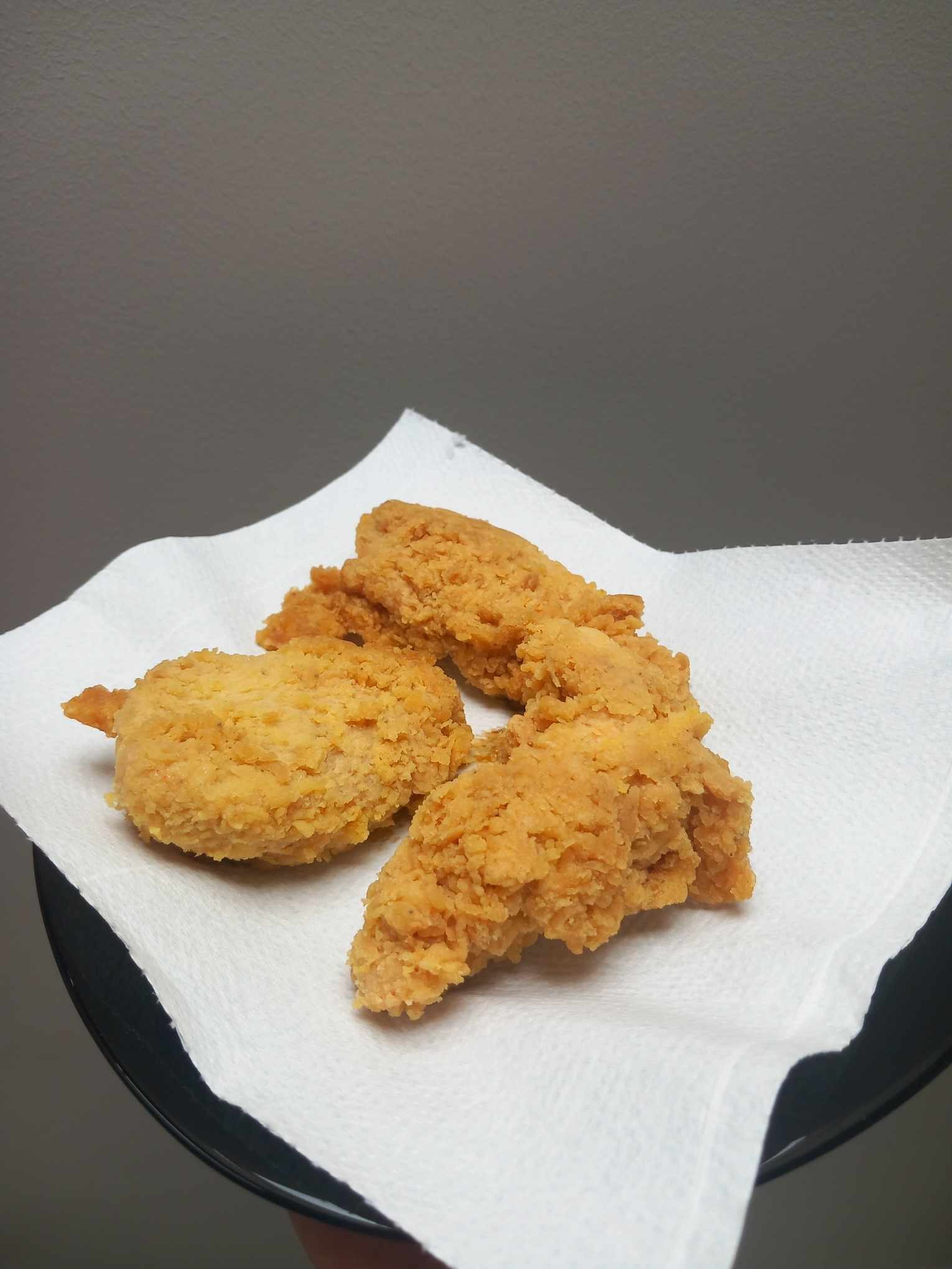 Chrupiące, złociste kawałki kurczaka w panierce na białej serwetce, ułożone na czarnym talerzu. Domowe jedzenie, ciepłe oświetlenie.