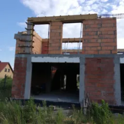 Surowy stan dwukondygnacyjnego budynku z pustaków ceramicznych, widoczne szalunki drewniane i podpora stropu na tle błękitnego nieba.