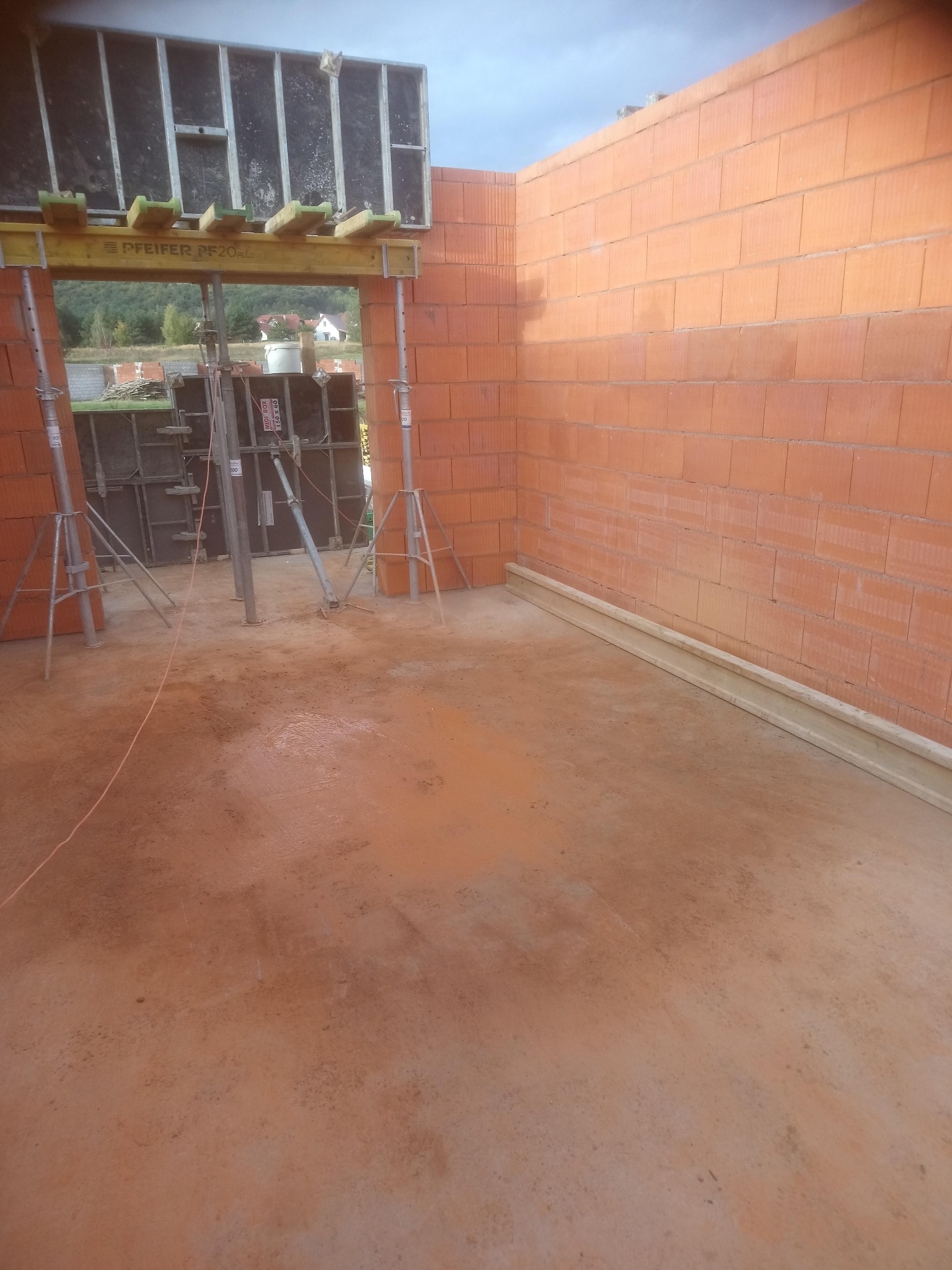 Wnętrze budynku w trakcie budowy z czerwonej cegły, widoczne szalunki nad otworem okiennym i podpory, podłoże ziemiste, w tle zielony krajobraz.