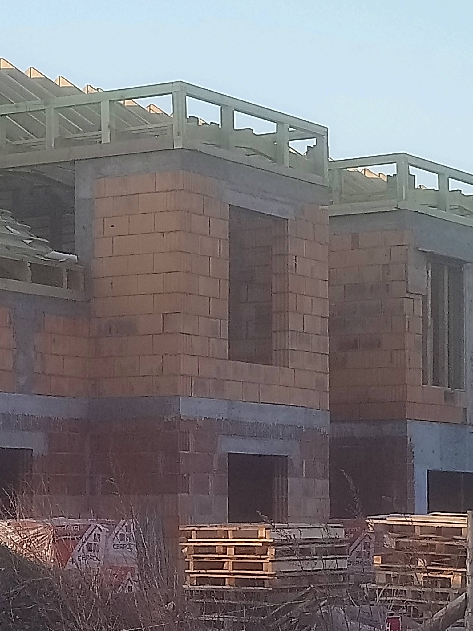 Wznoszenie ścian z cegły ceramicznej Porotherm na budowie domu dwukondygnacyjnego, widoczne palety z materiałem i konstrukcja dachu w trakcie montażu.