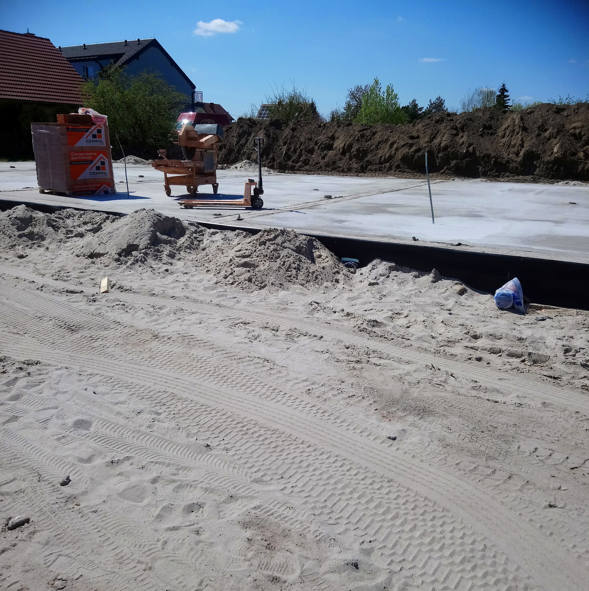 Przygotowany teren pod budowę domu: wylany fundament, obok paleta z cegłami ceramicznymi CERPOL i wózek paletowy, w tle pryzma ziemi.