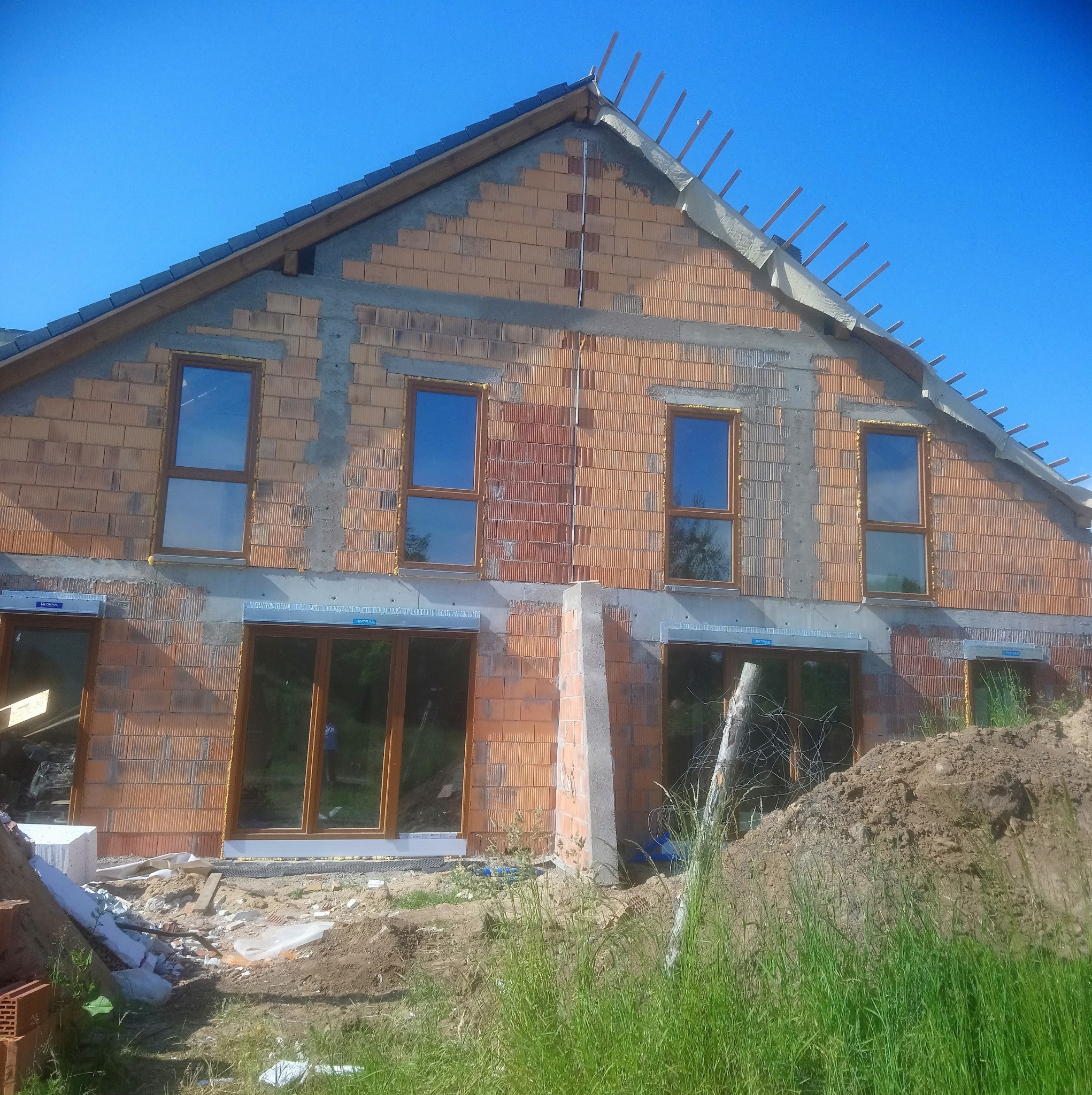 Surowy stan dwukondygnacyjnego budynku z czerwonej cegły z drewnianymi oknami, widoczne elementy konstrukcji dachu i nieotynkowane ściany zewnętrzne.