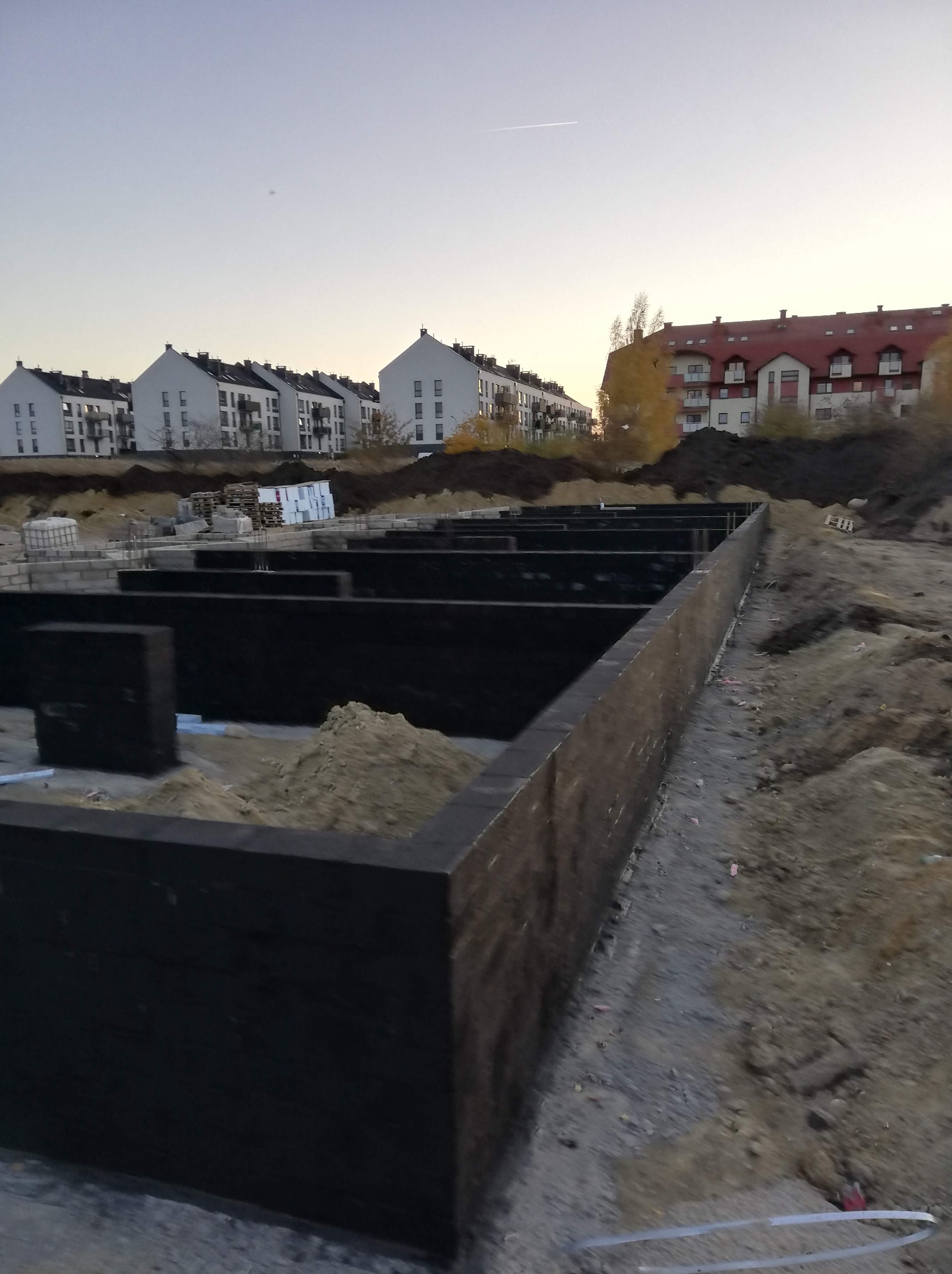 Przygotowany wykop pod fundamenty z zabezpieczonymi ścianami szalunkowymi, na tle zabudowy wielorodzinnej i wieczornego nieba z widocznym śladem po przelocie samolotu.