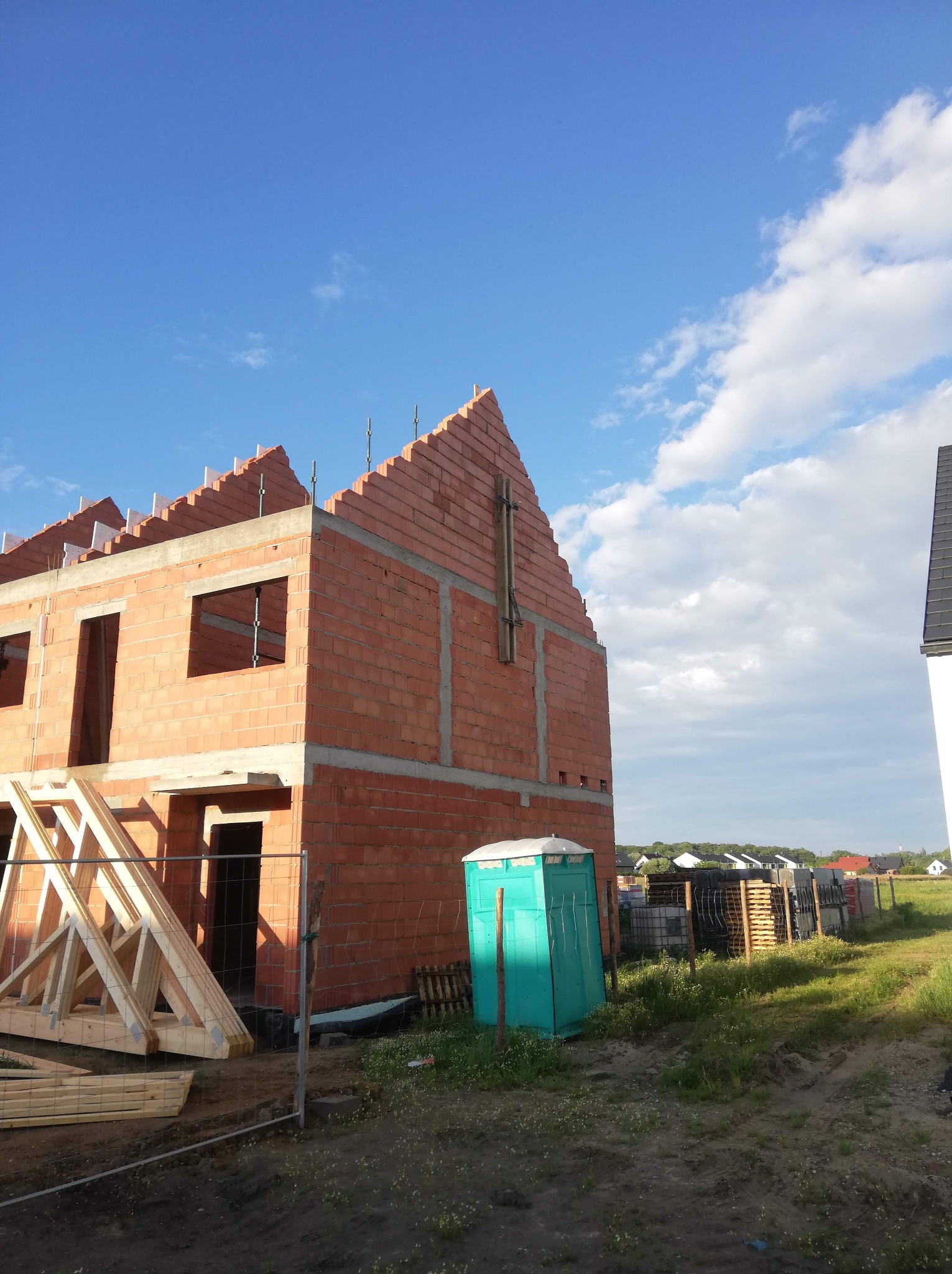 Wznoszenie dwukondygnacyjnego budynku z czerwonej cegły, widoczne niezamurowane otwory okienne i drzwiowe, z zamontowanymi elementami szalunkowymi na szczycie, na tle błękitnego nieba i okolicznej...