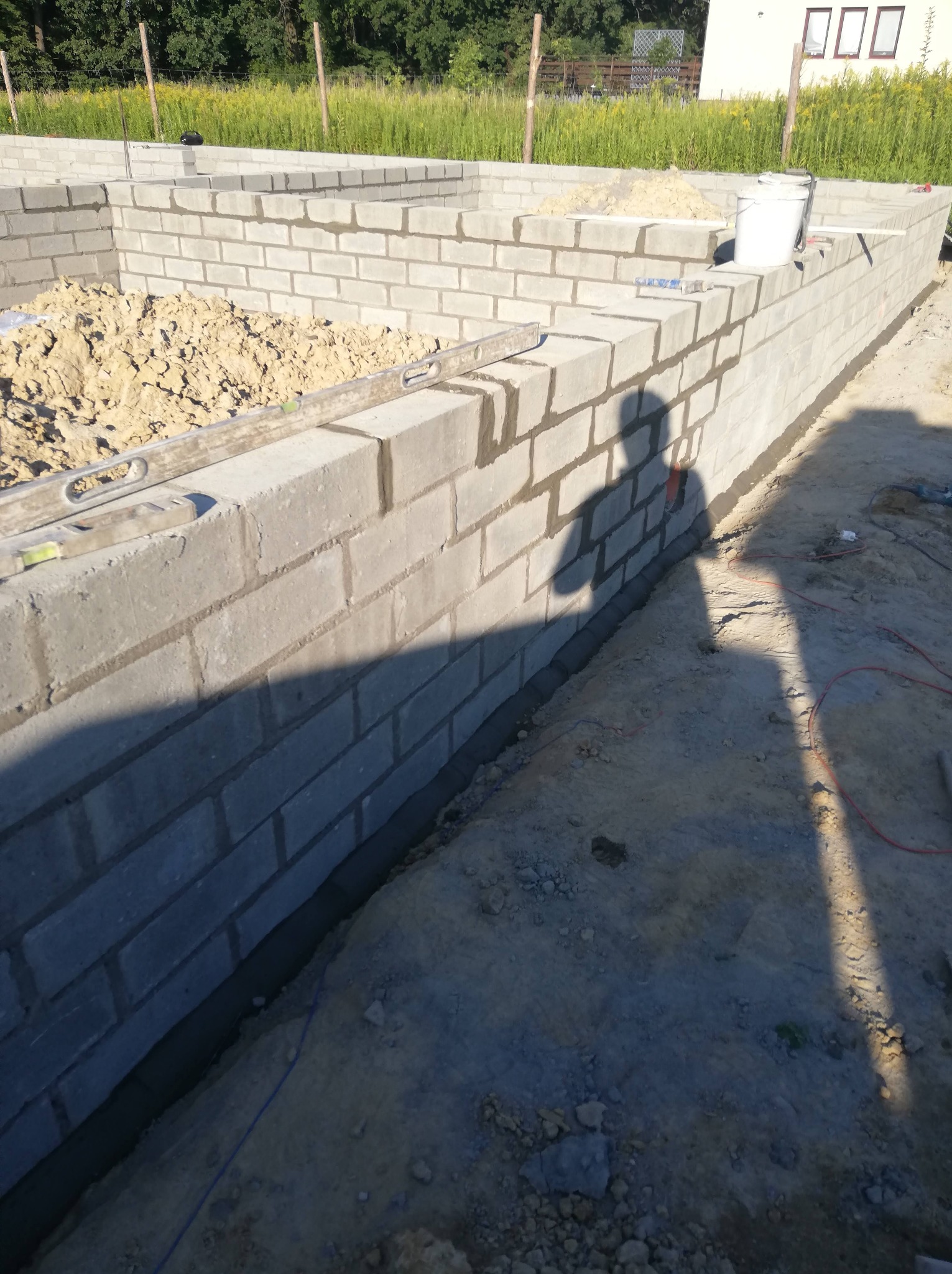 Wznoszenie fundamentów z bloczków betonowych, widoczny poziomica, cień osoby, teren budowy z piaskiem i roślinnością w tle.