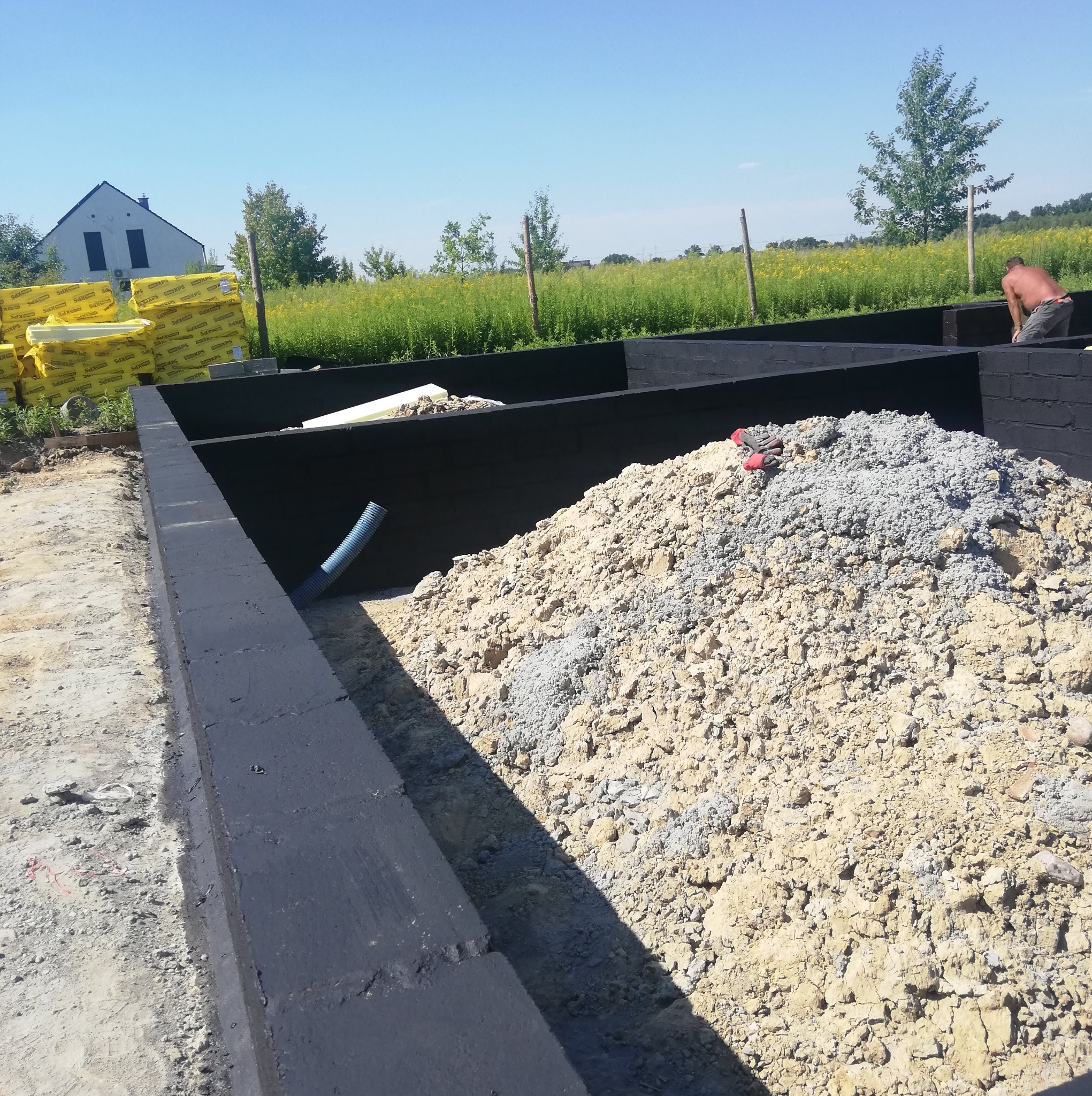 Przygotowanie fundamentów: zaizolowane ściany fundamentowe, stos izolacji termicznej oraz pryzma piasku z cementem na placu budowy, w tle dom i łąka.