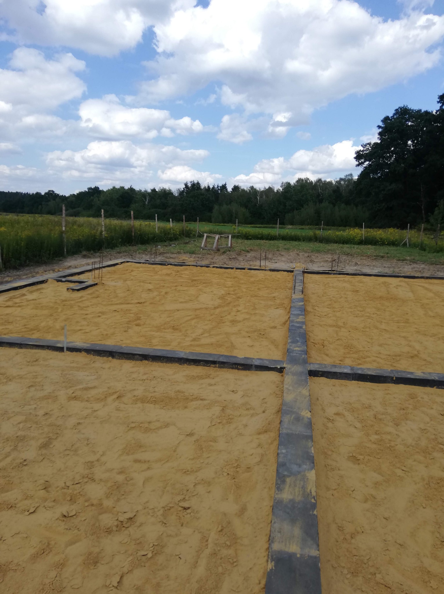 Przygotowany teren pod fundamenty z widocznymi szalunkami i wystającymi prętami zbrojeniowymi, tło stanowi łąka i las pod błękitnym niebem z chmurami.