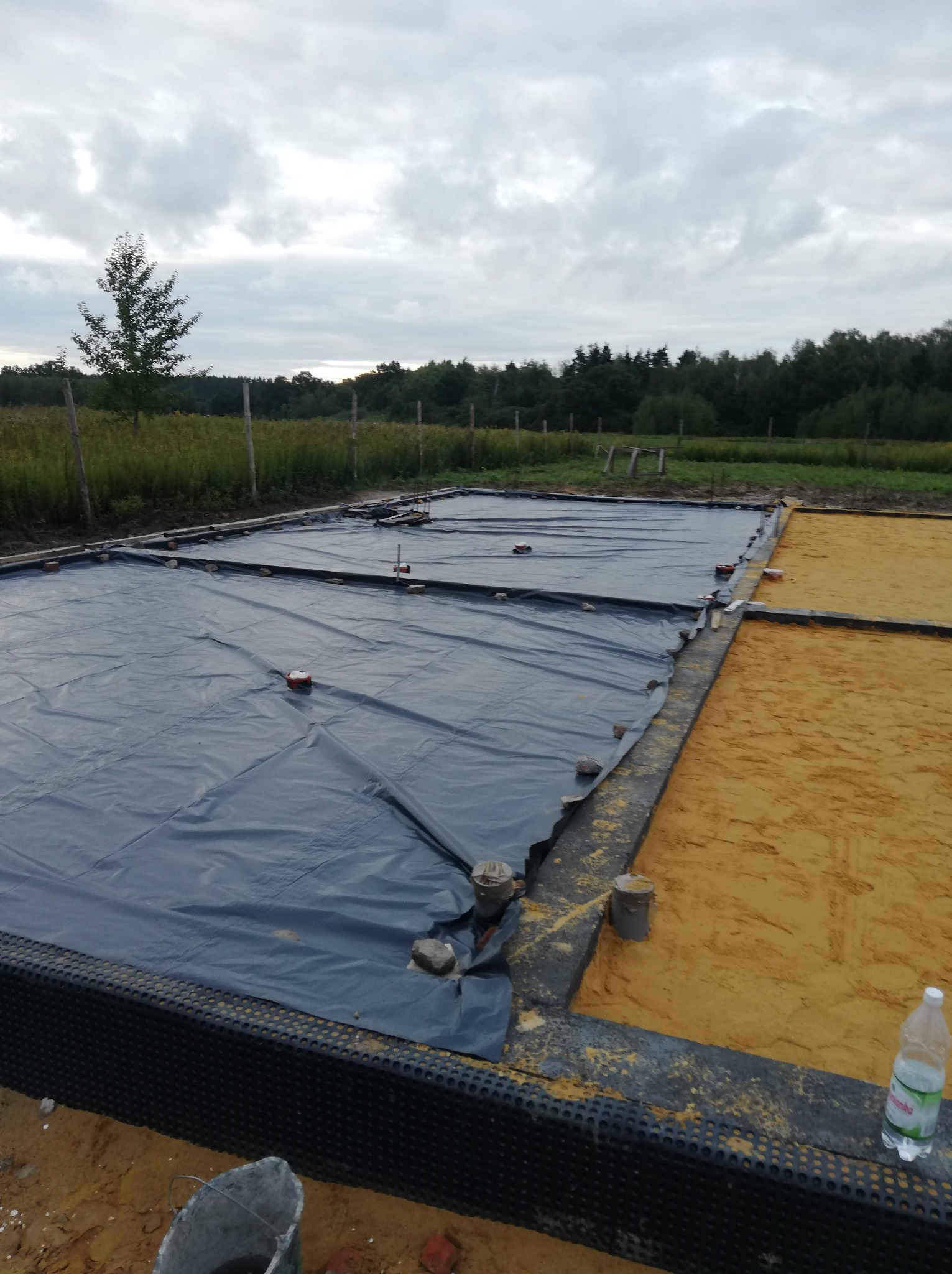 Przygotowany teren pod wylewkę fundamentów: czarna folia izolacyjna rozłożona na piasku, obrzeża zabezpieczone membraną kubełkową, widoczny kubeł i butelka wody.