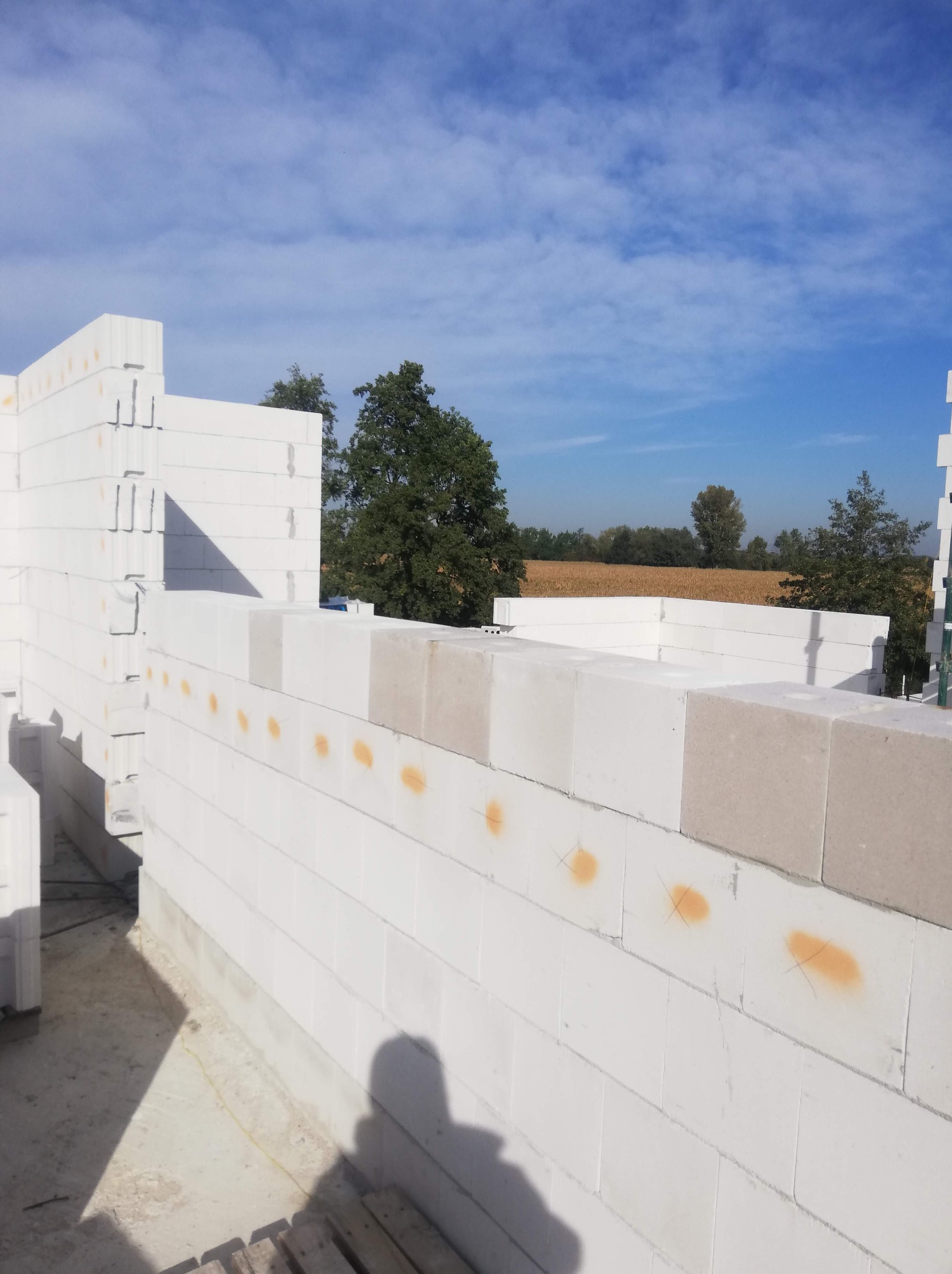 Wznoszenie ścian z białych bloczków, widoczne pomarańczowe oznaczenia na bloczkach, w tle pole i drzewa pod błękitnym niebem.