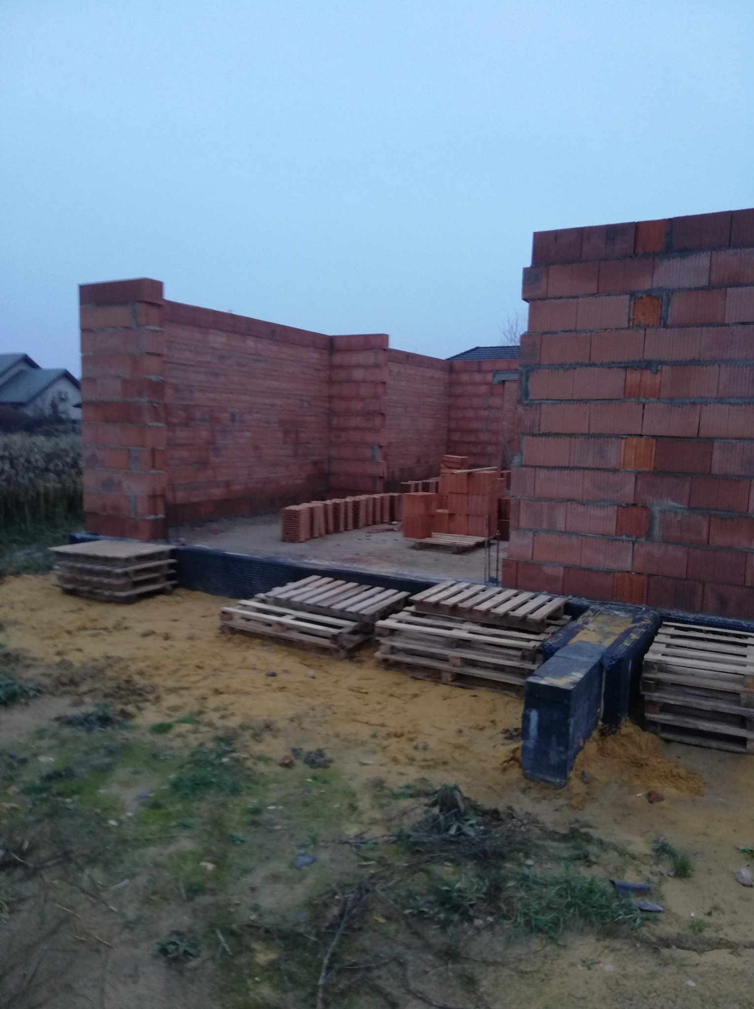 Wznoszone ściany parteru domu z pustaków ceramicznych na fundamencie z izolacją, palety z materiałami budowlanymi na placu budowy przy pochmurnym niebie.
