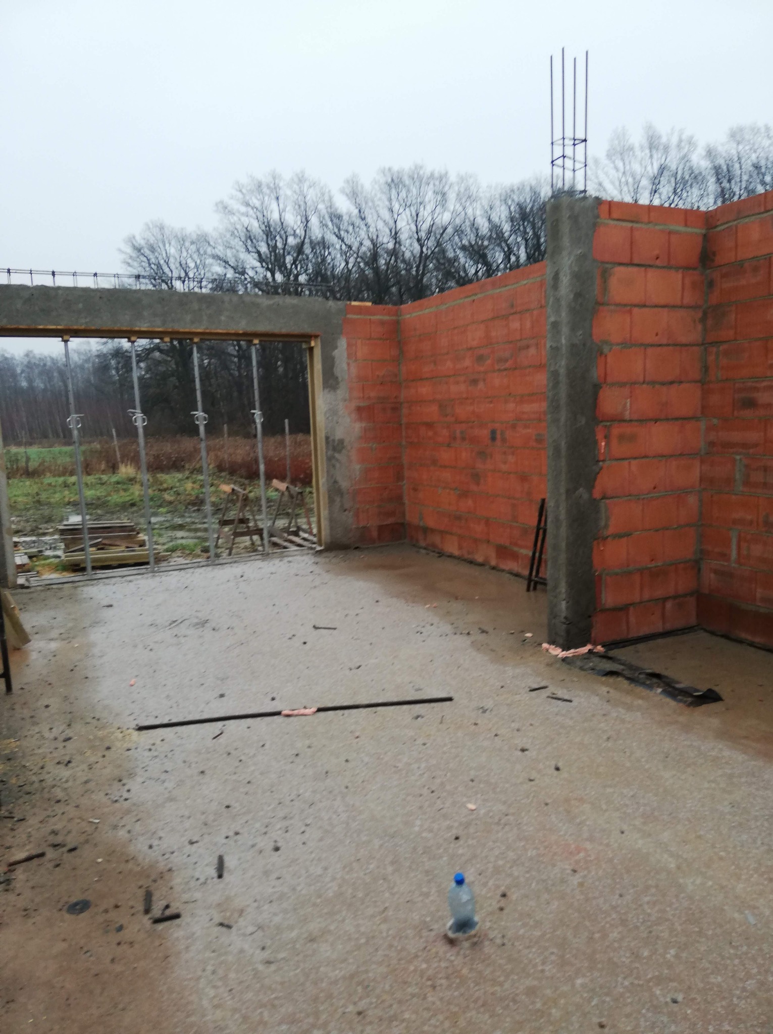 Wnętrze w trakcie budowy z widocznymi ścianami z czerwonej cegły, betonowym filarem z wystającymi prętami zbrojeniowymi, metalowymi podporami stropu i butelką wody na podłodze.