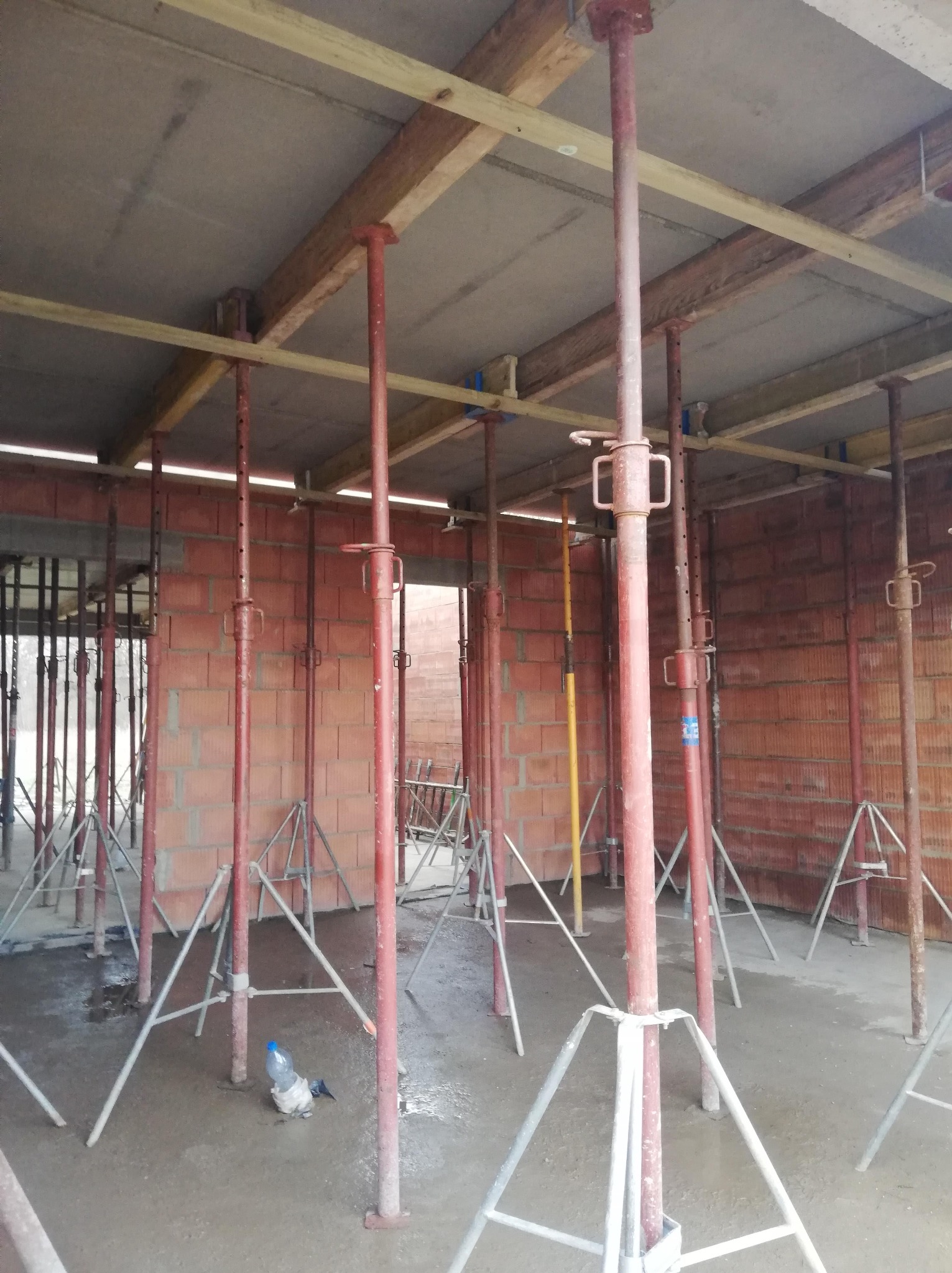 Wnętrze w trakcie budowy domu: widoczne czerwone ściany z pustaków, stalowe podpory stropu i drewniane belki. Na podłodze leży butelka z wodą.