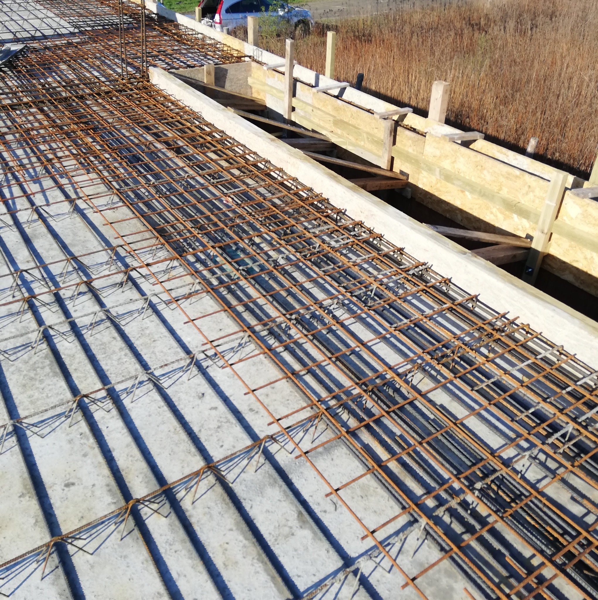 Zbrojenie płyty fundamentowej ułożone w szalunku z desek, widoczne z góry w słoneczny dzień.