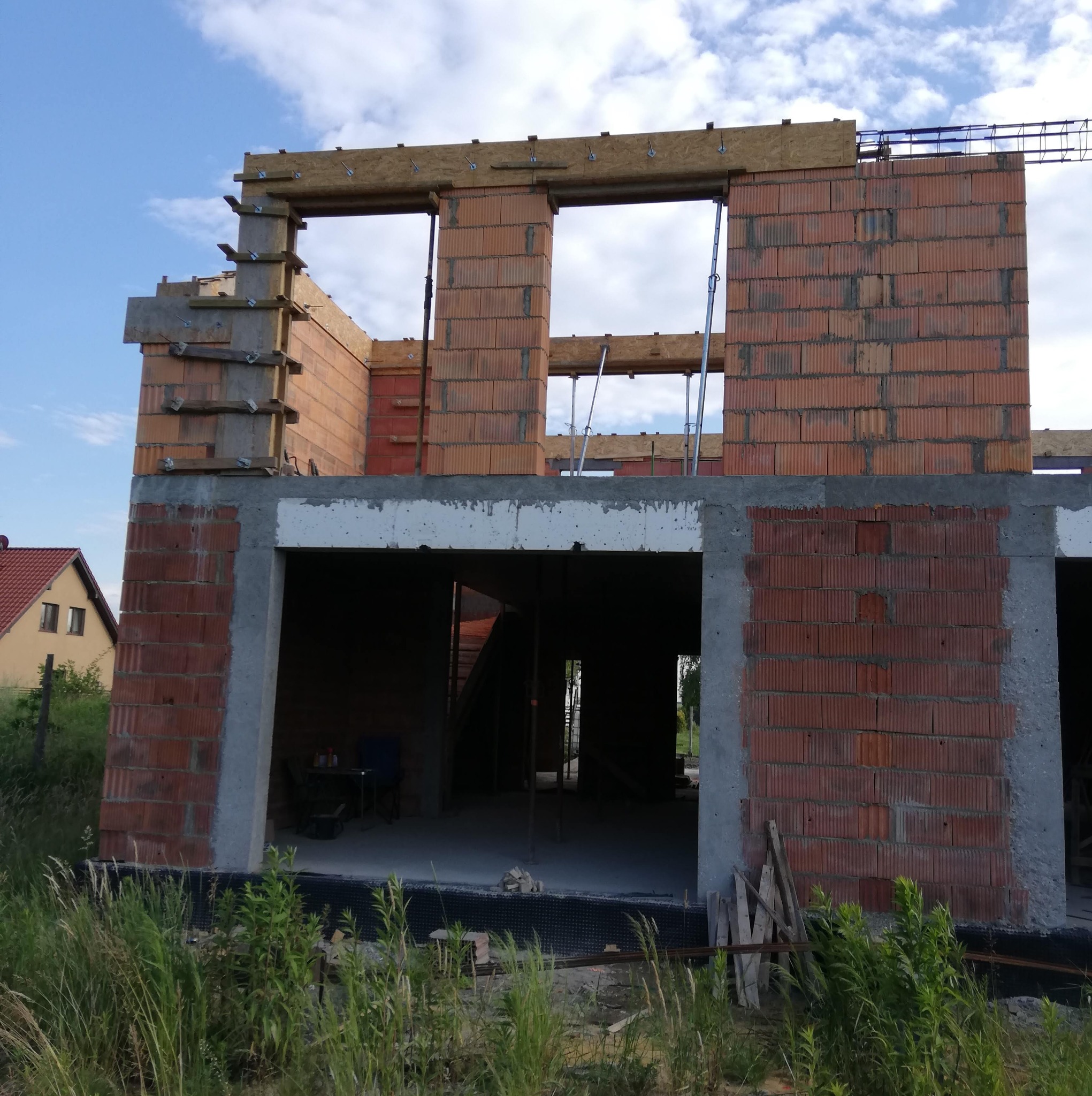 Surowy stan dwukondygnacyjnego budynku z pustaków ceramicznych, widoczne szalunki drewniane i podpora stropu na tle błękitnego nieba.