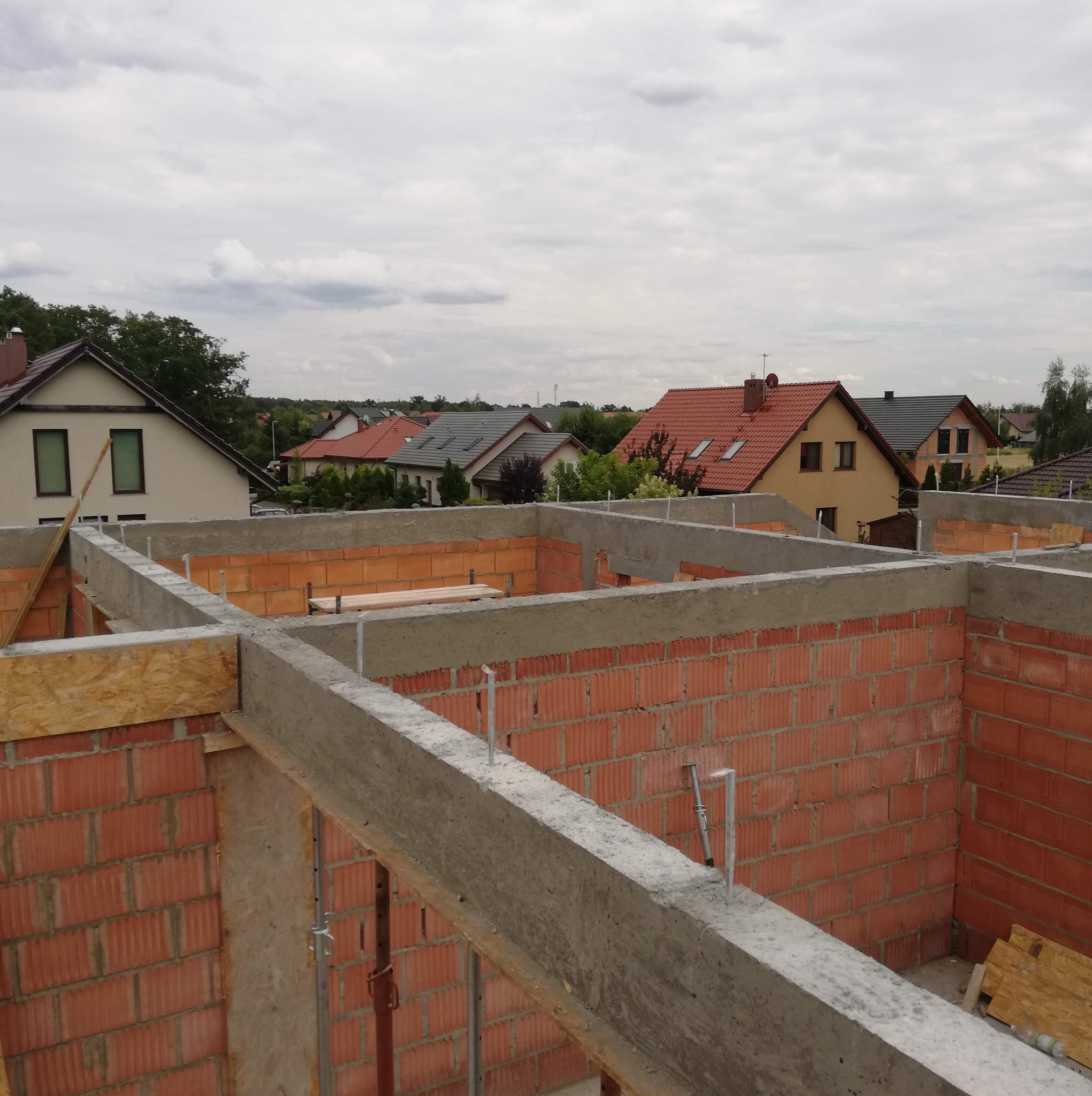 Wznoszenie ścian z czerwonej cegły z widocznymi betonowymi nadprożami i drewnianym szalunkiem na tle zabudowy jednorodzinnej pod pochmurnym niebem.