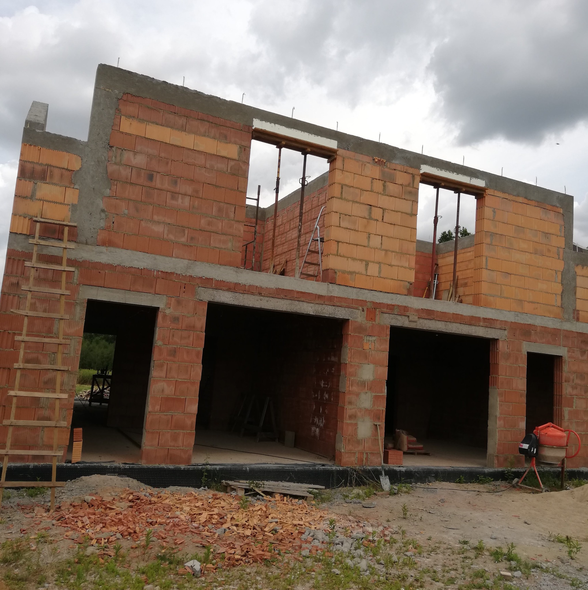 Dwukondygnacyjny budynek w trakcie budowy z czerwonej cegły ceramicznej, widoczne otwory okienne i drzwiowe, drewniane rusztowanie i drabina, gruz ceglany na ziemi, betoniarka obok budynku.