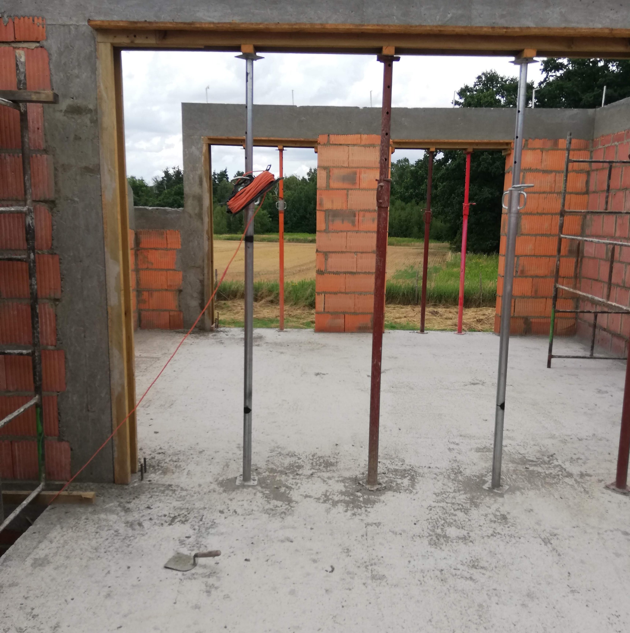 Wnętrze domu w trakcie budowy: surowe ściany z czerwonej cegły, betonowa podłoga, stalowe podpory, widok na zewnątrz przez otwory okienne, kielnia leżąca na podłodze, pomarańczowy zwój kabli...