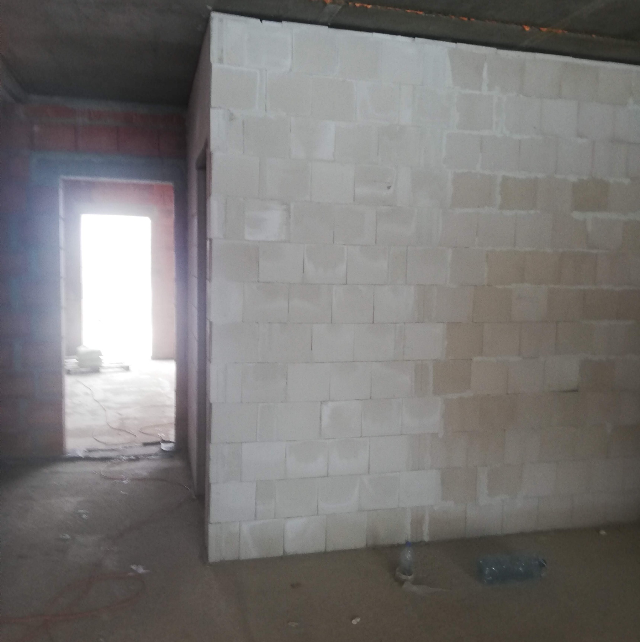 Wnętrze surowego domu w trakcie budowy, ściana z białych bloczków częściowo otynkowana, widoczne przejście do innego pomieszczenia z ceglanymi ścianami, betonowy strop i podłoga.