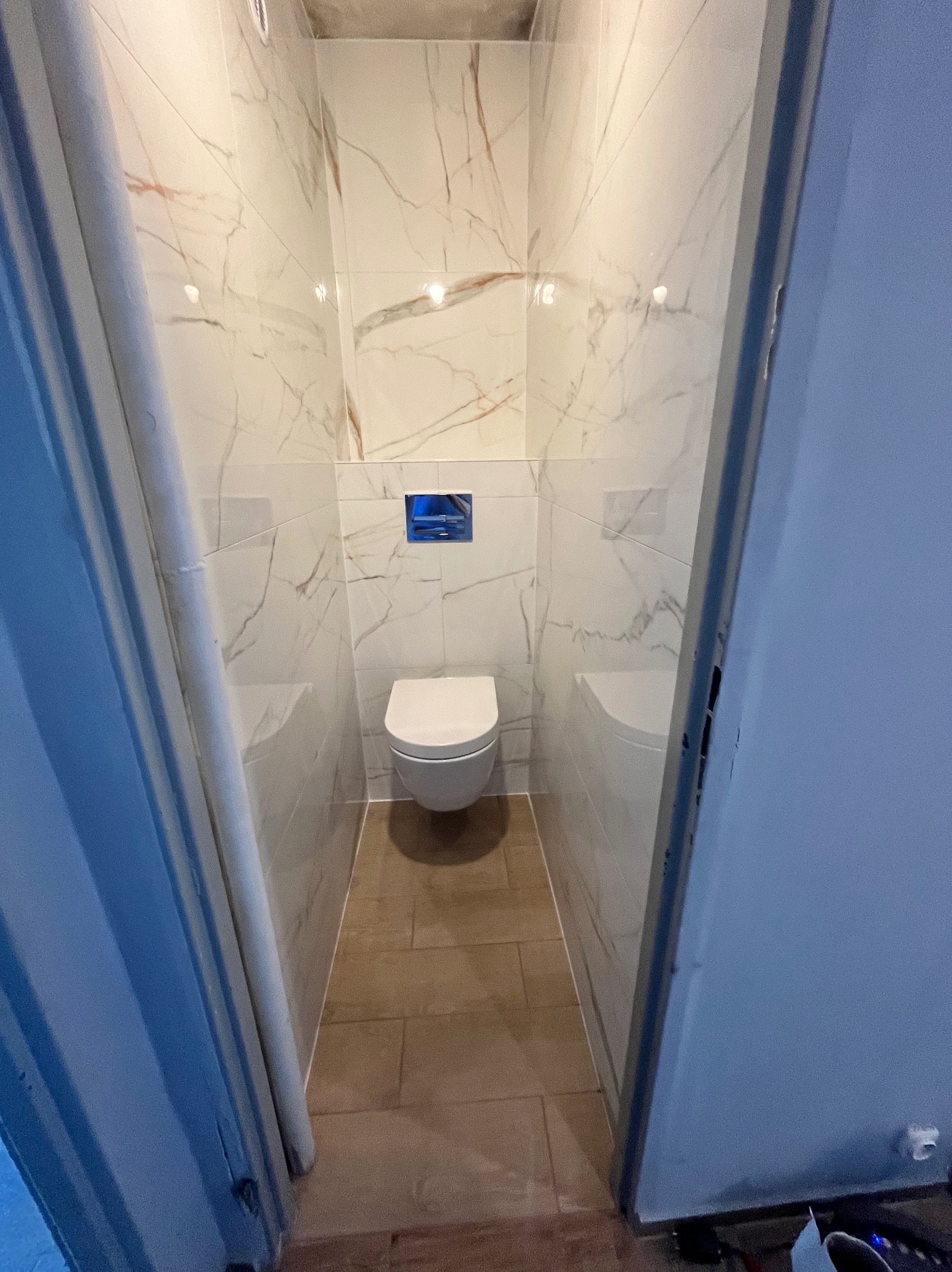 Wąska toaleta po remoncie z marmurowymi płytkami ściennymi i brązowymi podłogowymi, widoczna zza uchylonych drzwi. Niebieski przycisk spłuczki.