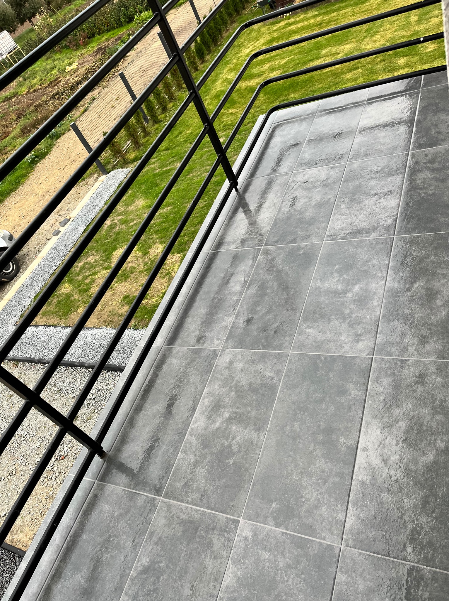 Błyszczące, szare płytki na balkonie z czarną, metalową balustradą. Widok z góry na świeżo ułożoną podłogę z płytek, zieleń trawnika w tle.