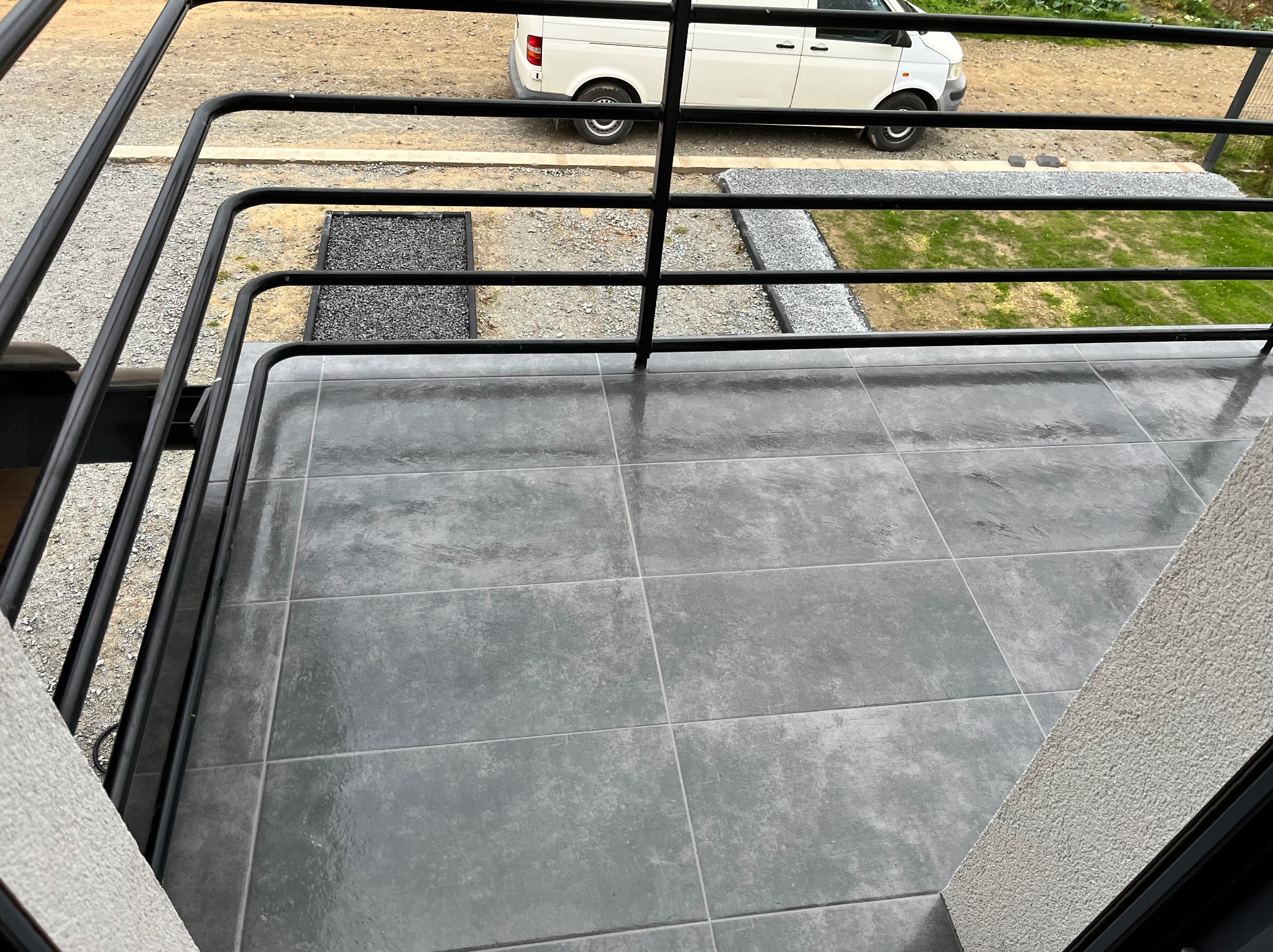 Balkon z szarymi płytkami, czarną balustradą i widokiem na podwórze z białym samochodem dostawczym w tle. Minimalistyczny design.