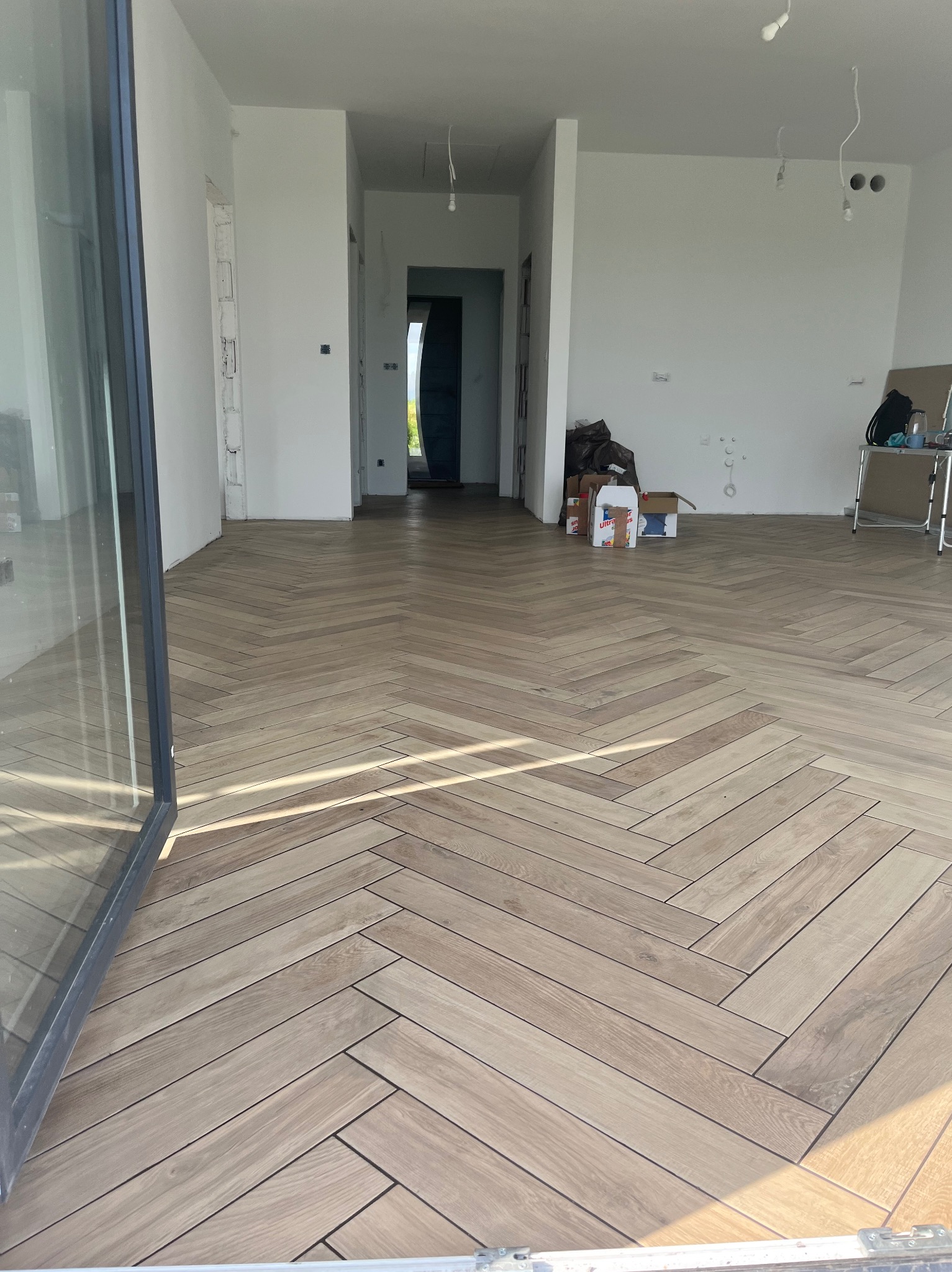 Ułożona podłoga z paneli w jodełkę w jasnym, przestronnym wnętrzu. Widoczne niedokończone ściany i otwarte przejścia do innych pomieszczeń. W tle kartony i drabina.