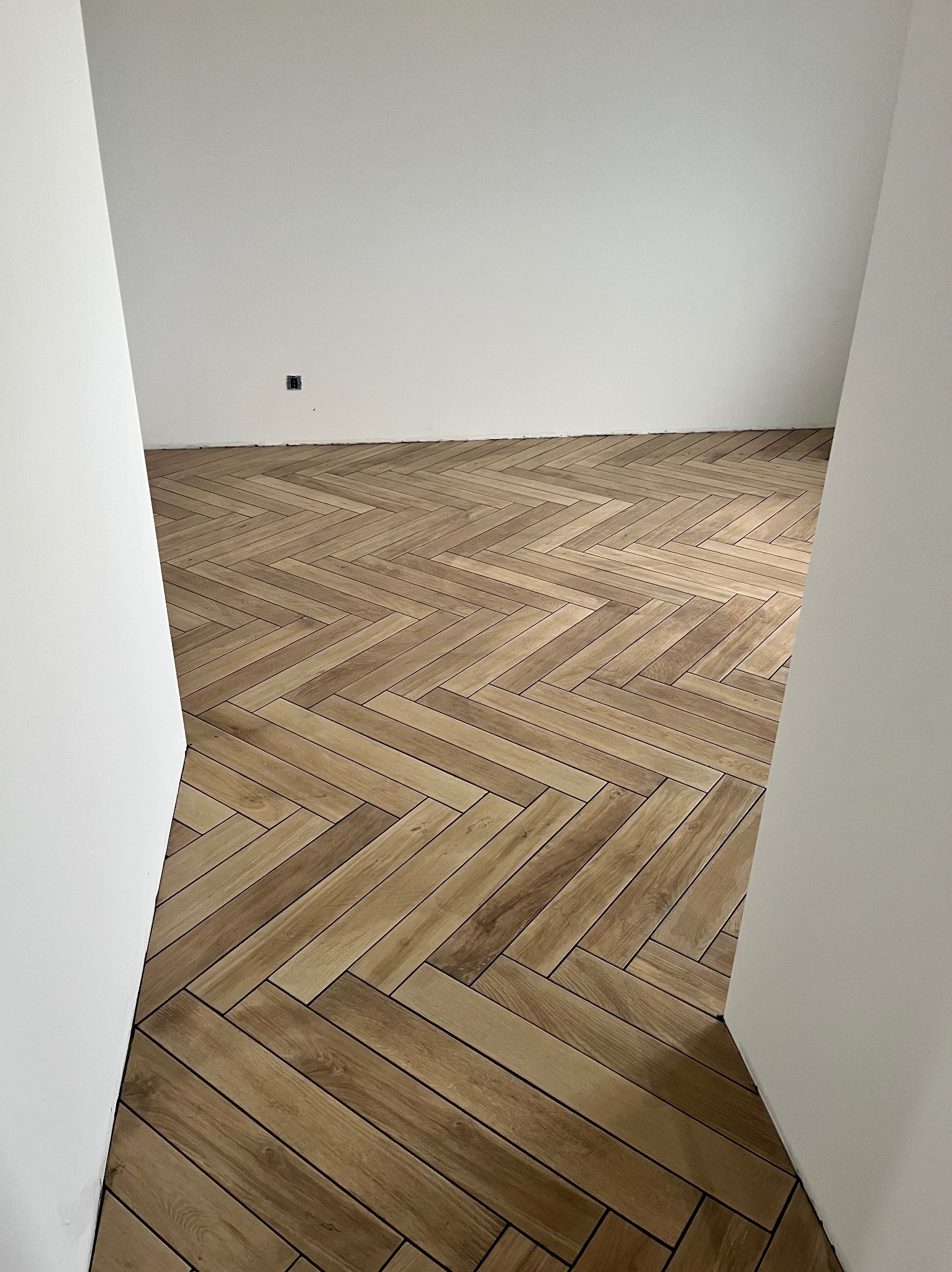 Ukończona podłoga z płytek imitujących drewno, ułożonych w jodełkę, widoczna z perspektywy wejścia do pomieszczenia. Białe ściany i gniazdko elektryczne.