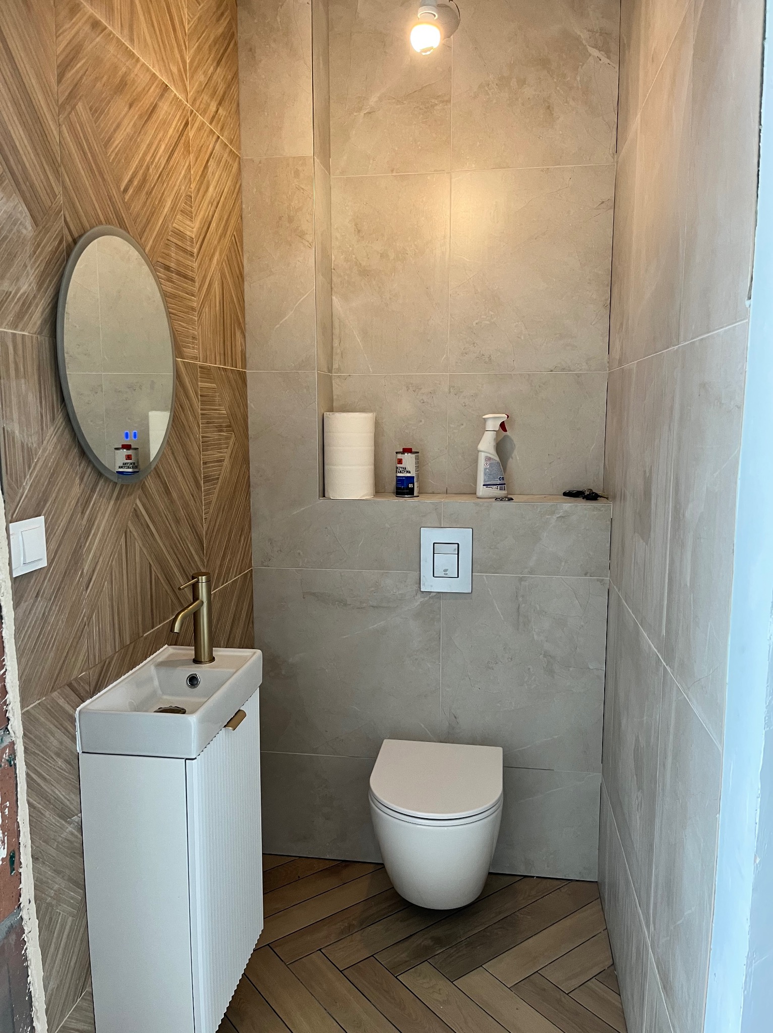 Nowoczesna łazienka z beżowymi płytkami, drewnopodobną podłogą w jodełkę i owalnym lustrem. Uwagę zwraca złota bateria umywalkowa i minimalistyczna toaleta.