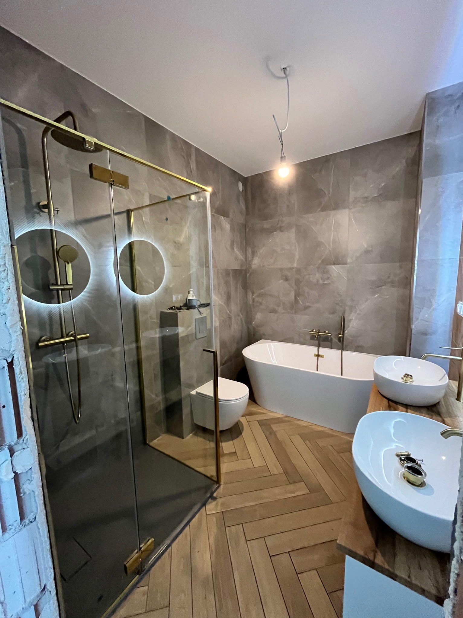 Nowoczesna łazienka z kabiną prysznicową typu walk-in z złotymi detalami, wolnostojącą wanną i dwiema umywalkami nablatowymi. Parkiet ułożony w jodełkę.