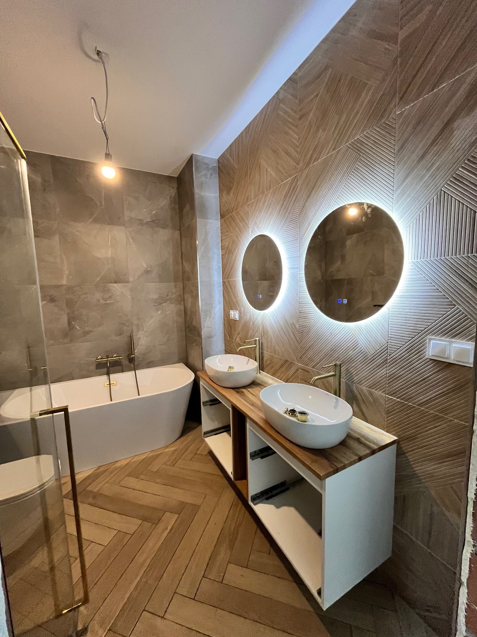Nowoczesna łazienka z drewnianą podłogą w jodełkę, owalną wanną, umywalkami na blacie i podświetlanymi lustrami na dekoracyjnej ścianie z płytek.