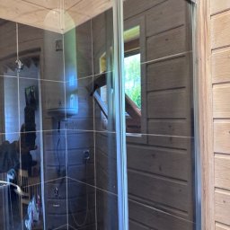 Porządkowa Symfonia - Szklana kabina prysznicowa w drewnianej łazience z widocznym oknem i bojlerem. Wnętrze wykończone drewnem, widoczna armatura.