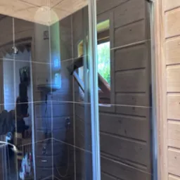 Szklana kabina prysznicowa w drewnianej łazience z widocznym oknem i bojlerem. Wnętrze wykończone drewnem, widoczna armatura.