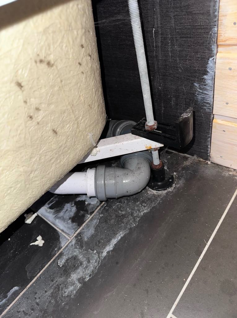 Niedokładnie umyta podłoga pod wanną z widocznymi rurami odpływowymi, zabrudzeniami i zaciekami. Niedoczyszczone fugi na ciemnych płytkach podłogowych.