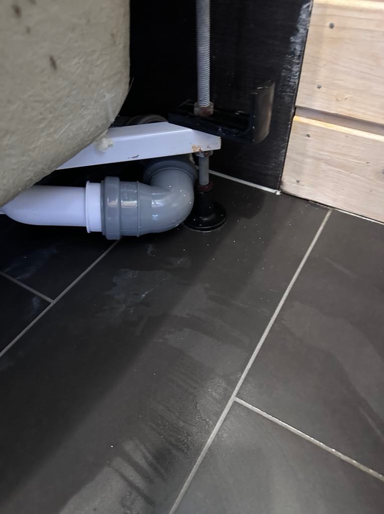 Szare płytki podłogowe, fragment odpływu wanny z szarą rurą, regulowana nóżka wanny. Widoczne ślady po sprzątaniu.