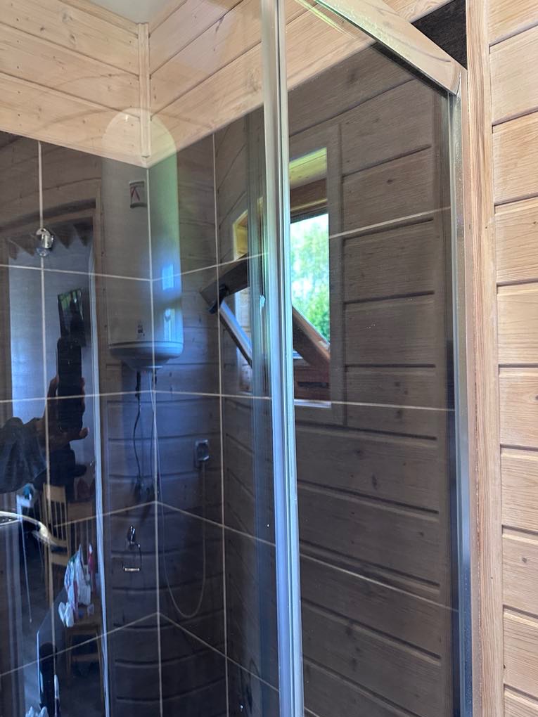 Szklana kabina prysznicowa w drewnianej łazience z widocznym oknem i bojlerem. Wnętrze wykończone drewnem, widoczna armatura.