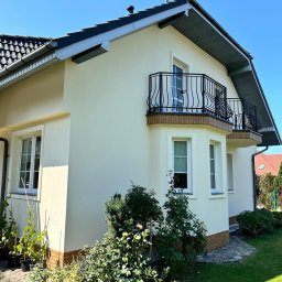 ELEWACJE-DACHY Słupsk - Elewacja jasnego domu jednorodzinnego z dwoma balkonami z ozdobnymi, czarnymi balustradami, widok z poziomu ogrodu z bujną roślinnością.