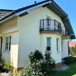 Elewacja jasnego domu jednorodzinnego z dwoma balkonami z ozdobnymi, czarnymi balustradami, widok z poziomu ogrodu z bujną roślinnością.